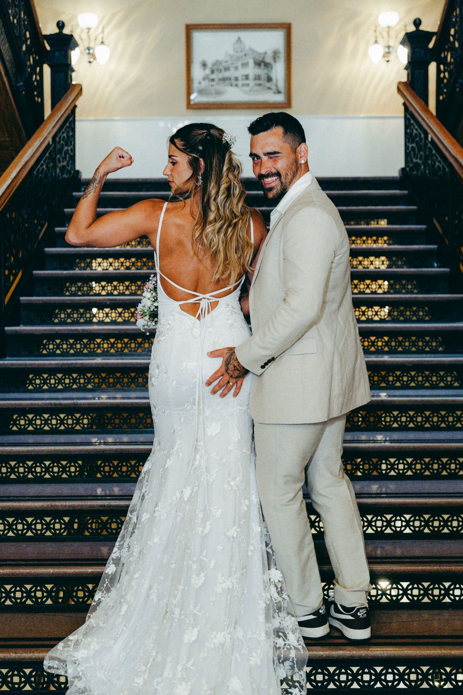 A couple poses playfully on elegant stairs with ornate railings in wedding attire.
