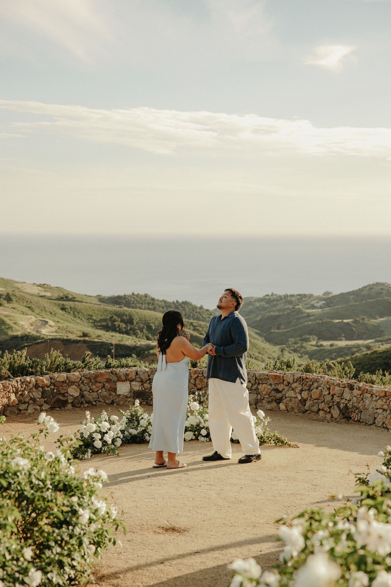 Couple holding hands at a scenic overlook with rolling green hills and ocean views, surrounded by white floral arrangements.