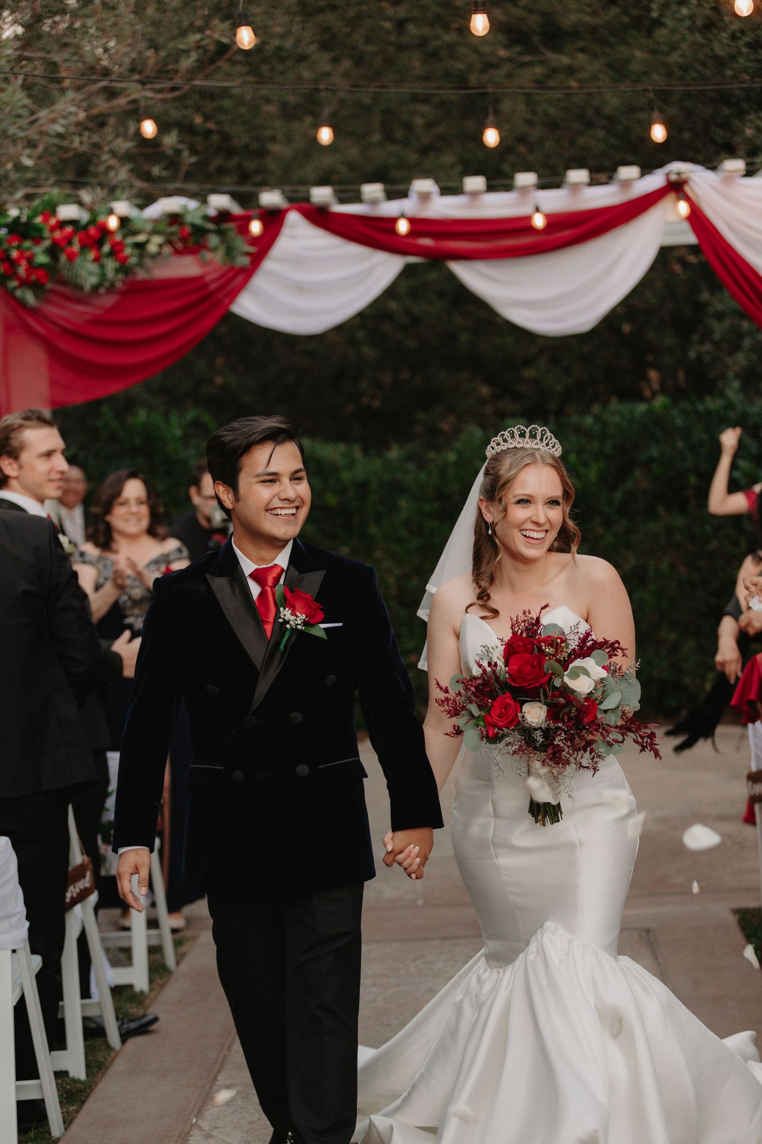 Wedding celebration outdoors with red and white decorations as couple walks down aisle under string lights and draped fabric.