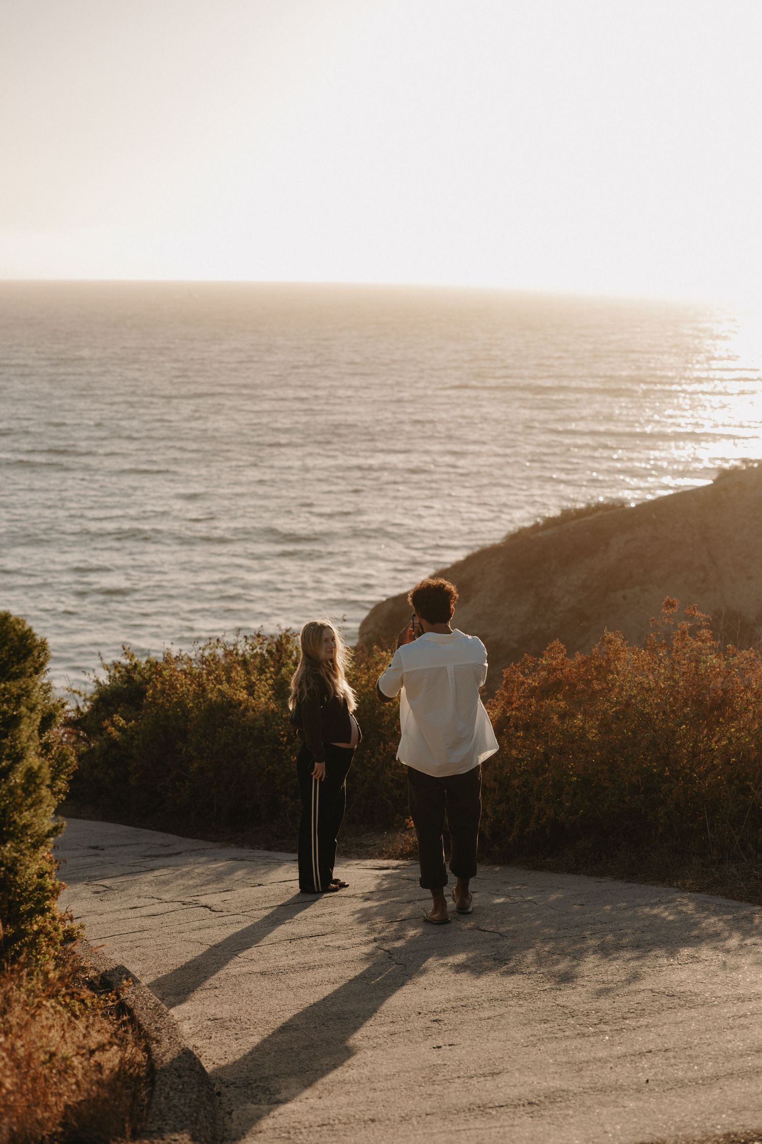 Two people walking along a coastal path at sunset with dramatic ocean views and golden light casting long shadows.
