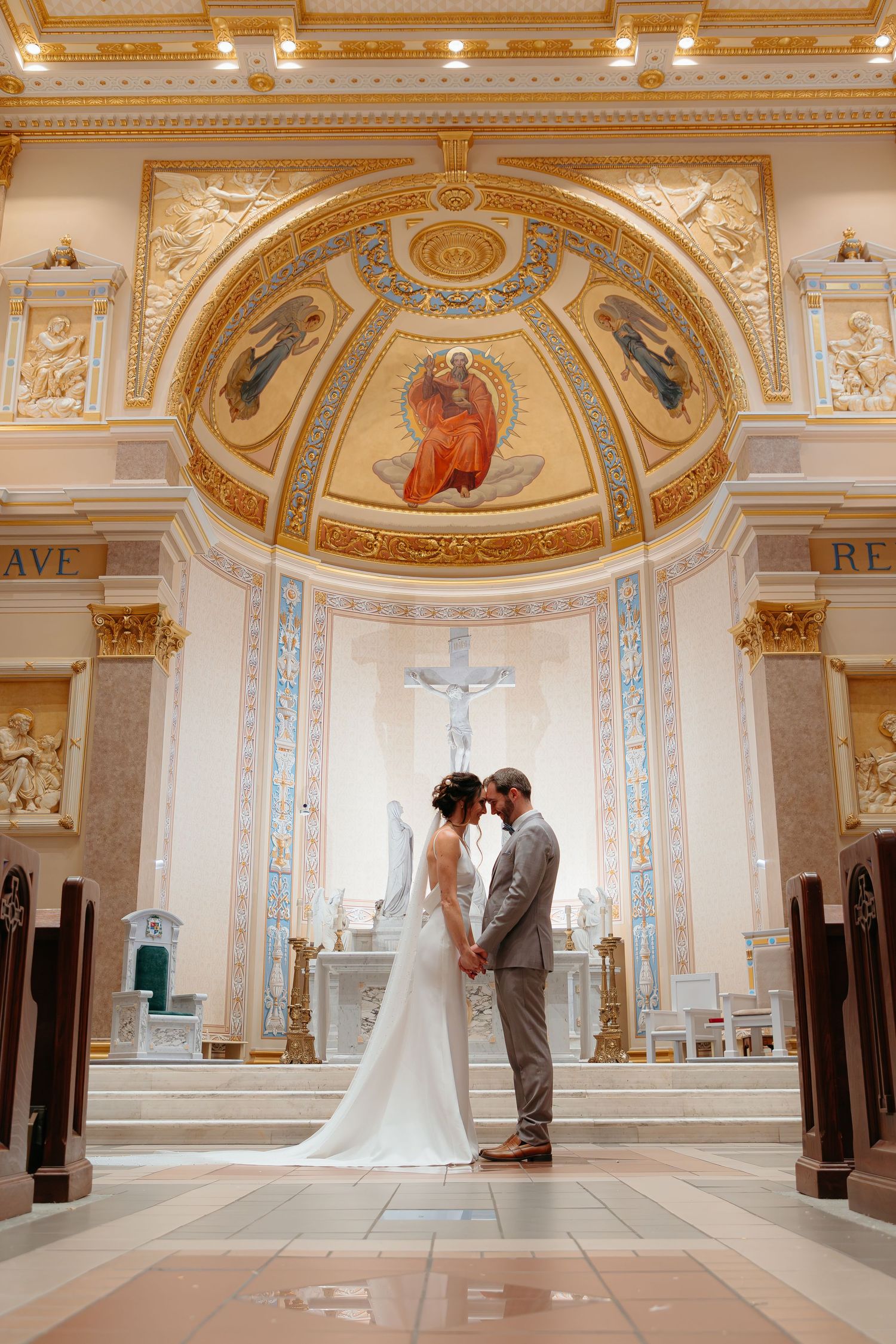 A wedding couple stands in elegant grand hall with ornate gold trim and arched ceilings.