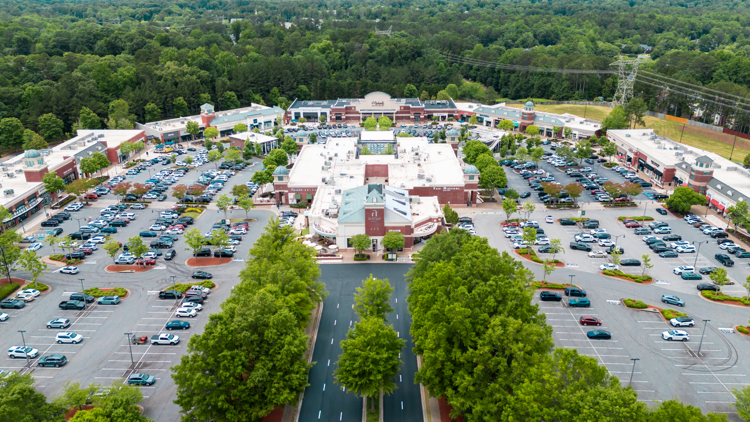 Aerial view of a busy shopping center parking lot surrounded by green trees and landscaping.