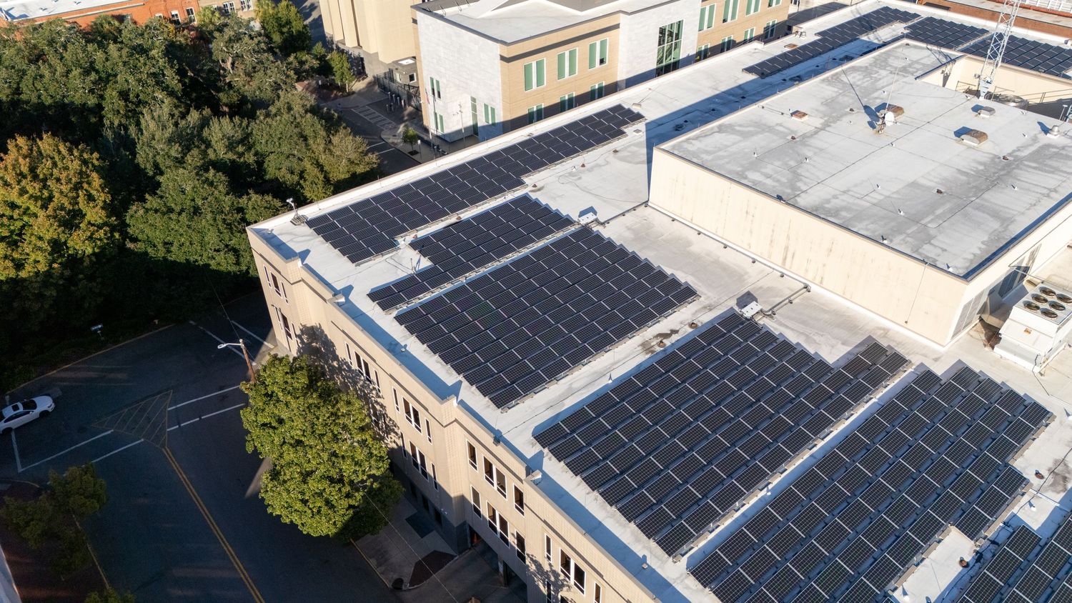Aerial view of commercial building rooftop with solar panel installation and surrounding trees in urban setting.