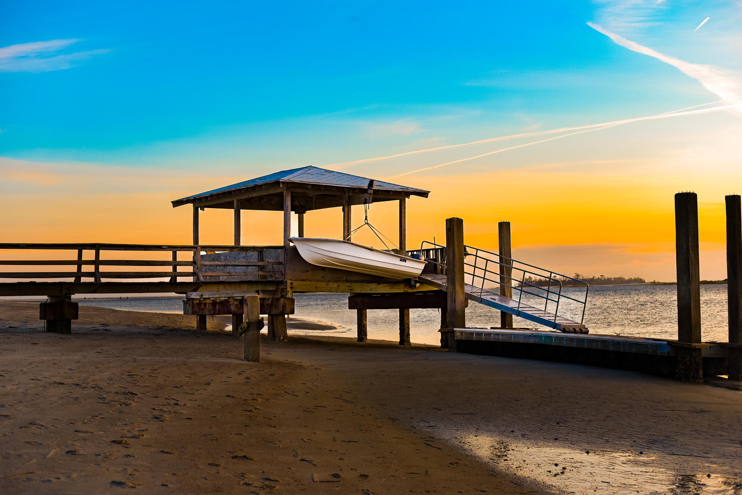 Wooden pier stretches into calm ocean waters at sunset with vibrant orange and blue sky.