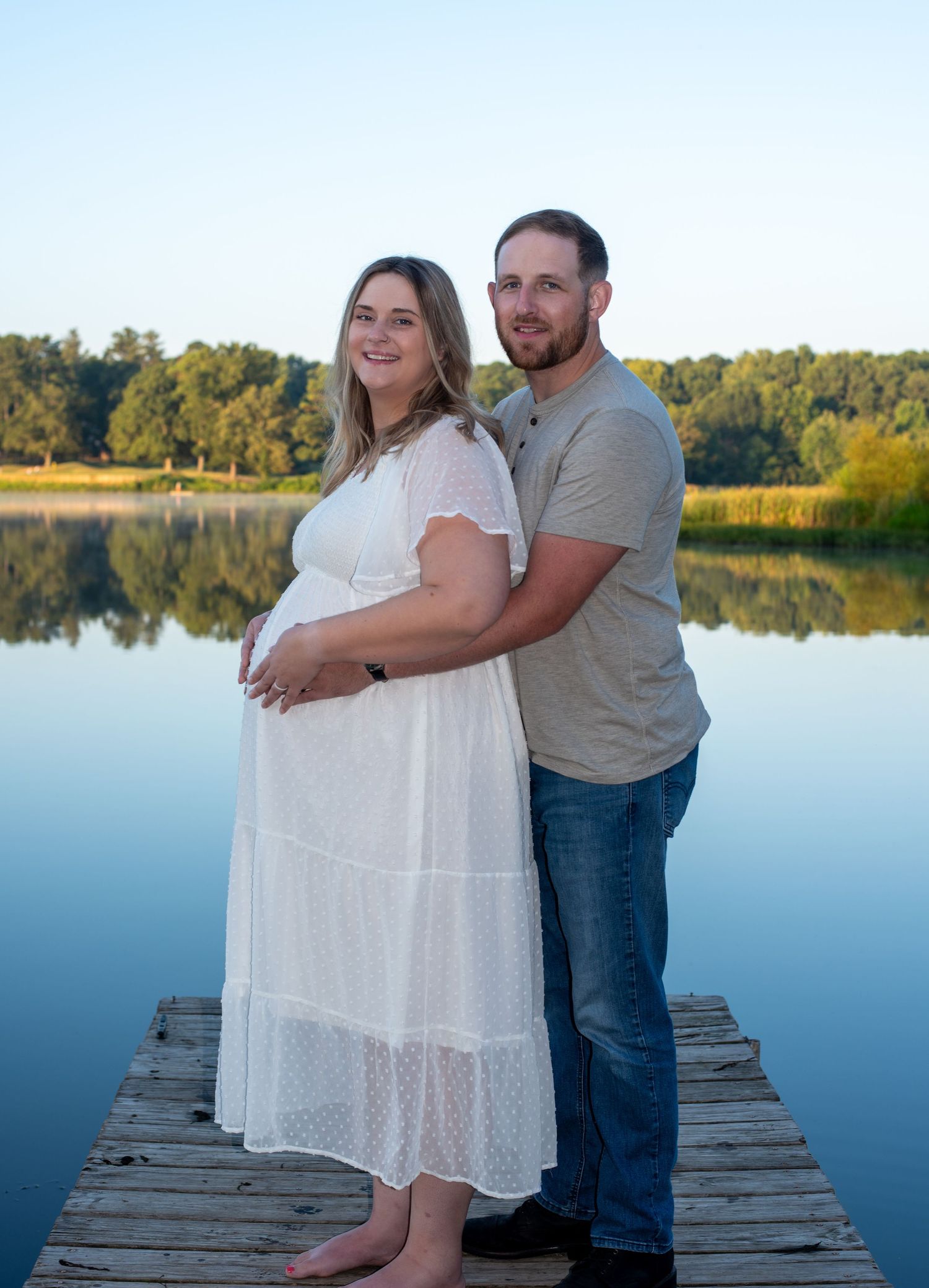 A couple stands together on a wooden dock by a calm lake during a maternity photo shoot at sunset.