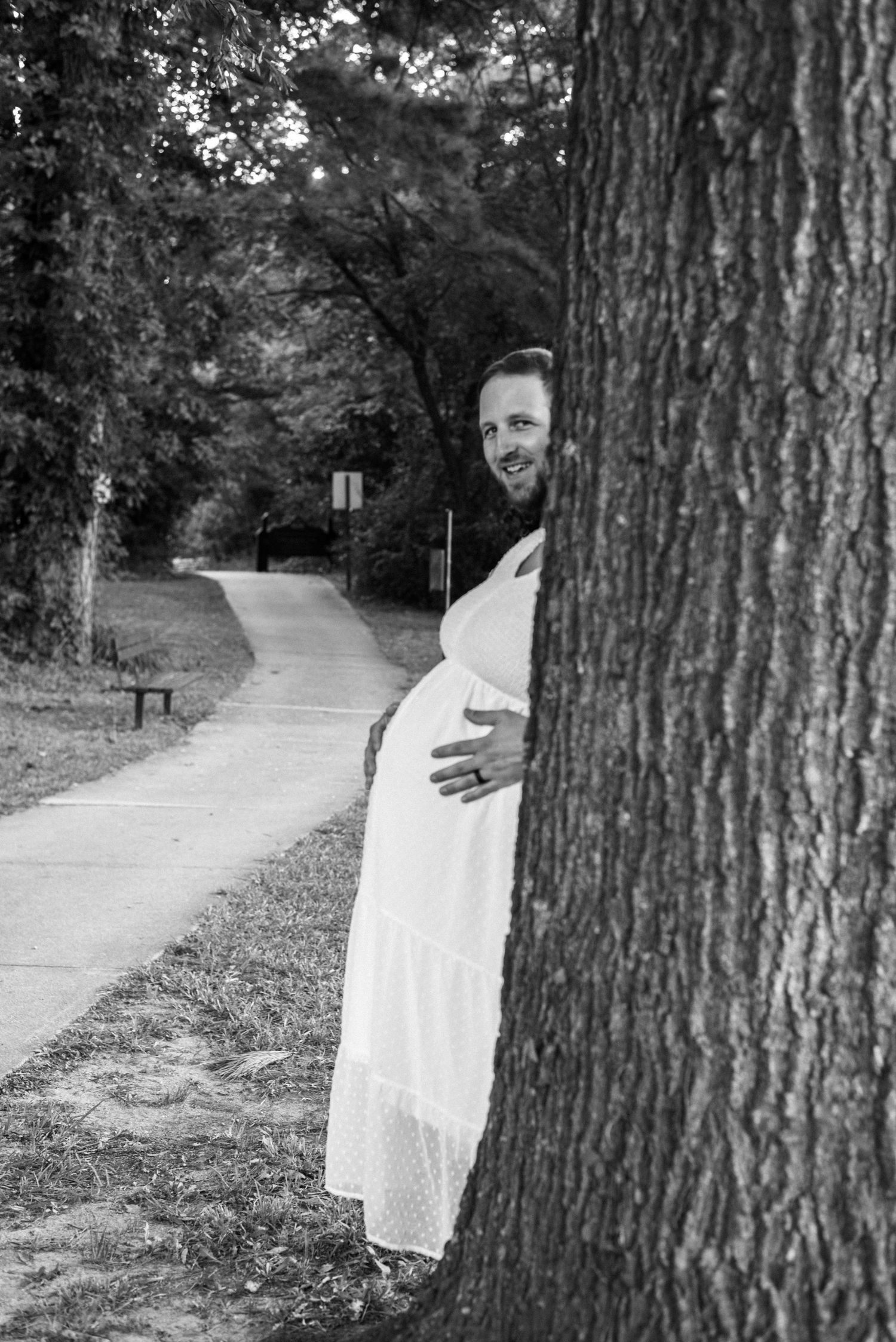 Humorous black and white maternity photos taken in a park setting with large trees.