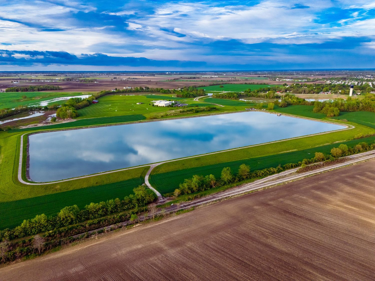 Aerial image of a water reservoir outside a city with green agricultural fields under a bright blue sky with white clouds.