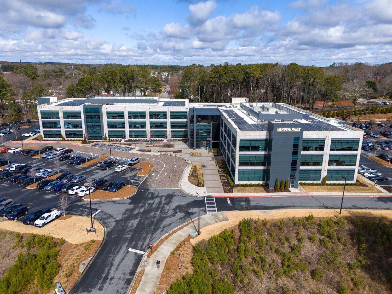 Aerial view of modern commercial building complex with parking lots and landscaping under blue sky.