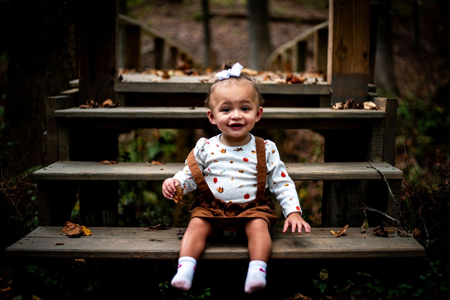 A child sits on a rustic wooden bench outdoors in casual clothing with a natural backdrop.