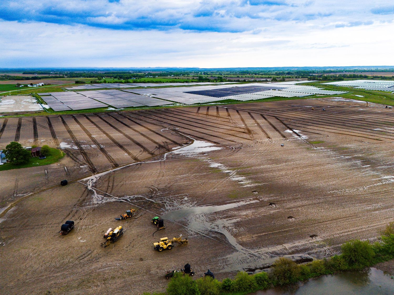 Aerial view of agricultural fields with harvesting equipment and irrigation systems under cloudy blue skies.