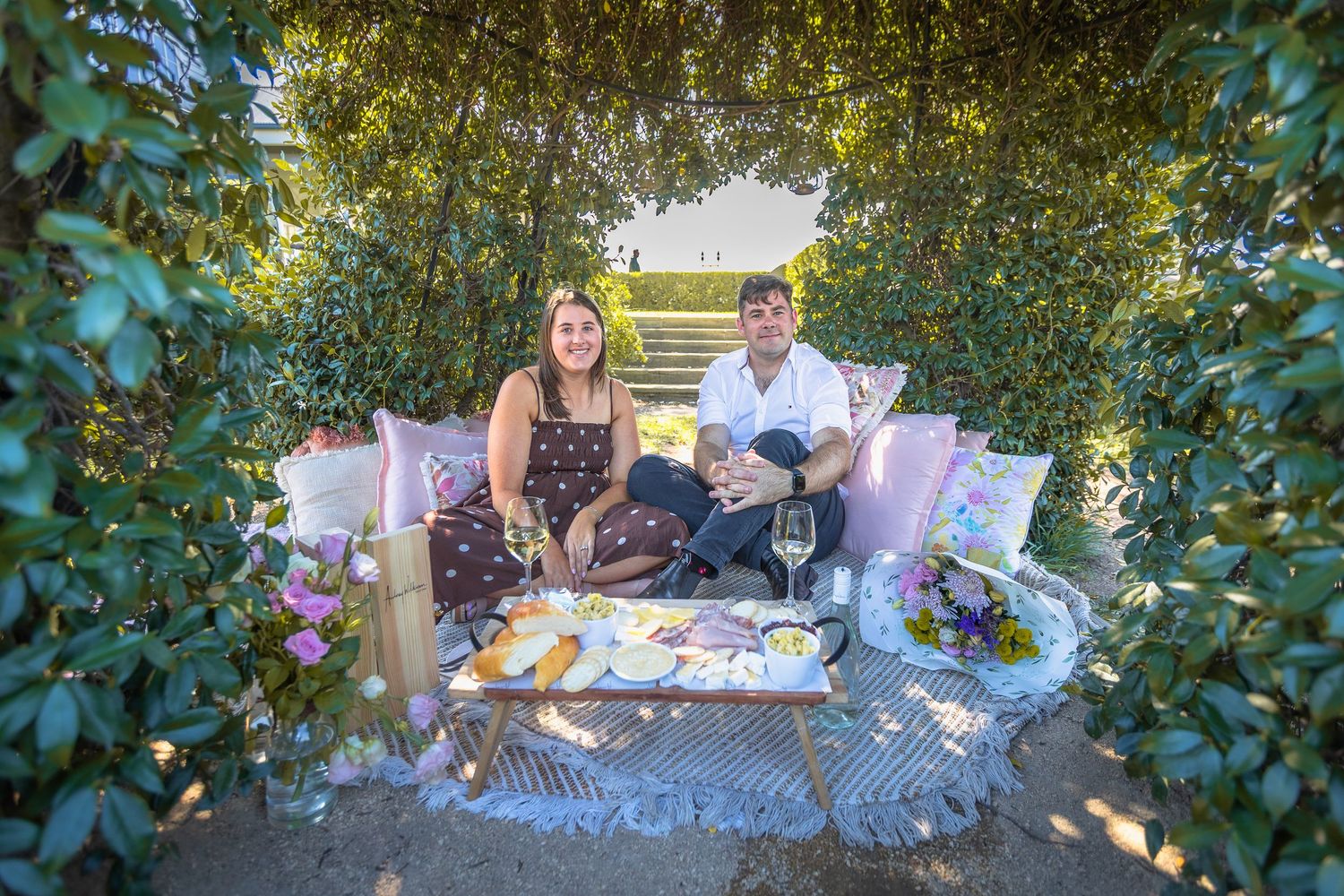 Couple enjoying a romantic outdoor picnic with wine, cheese, and cushions in a lush green garden arbor.