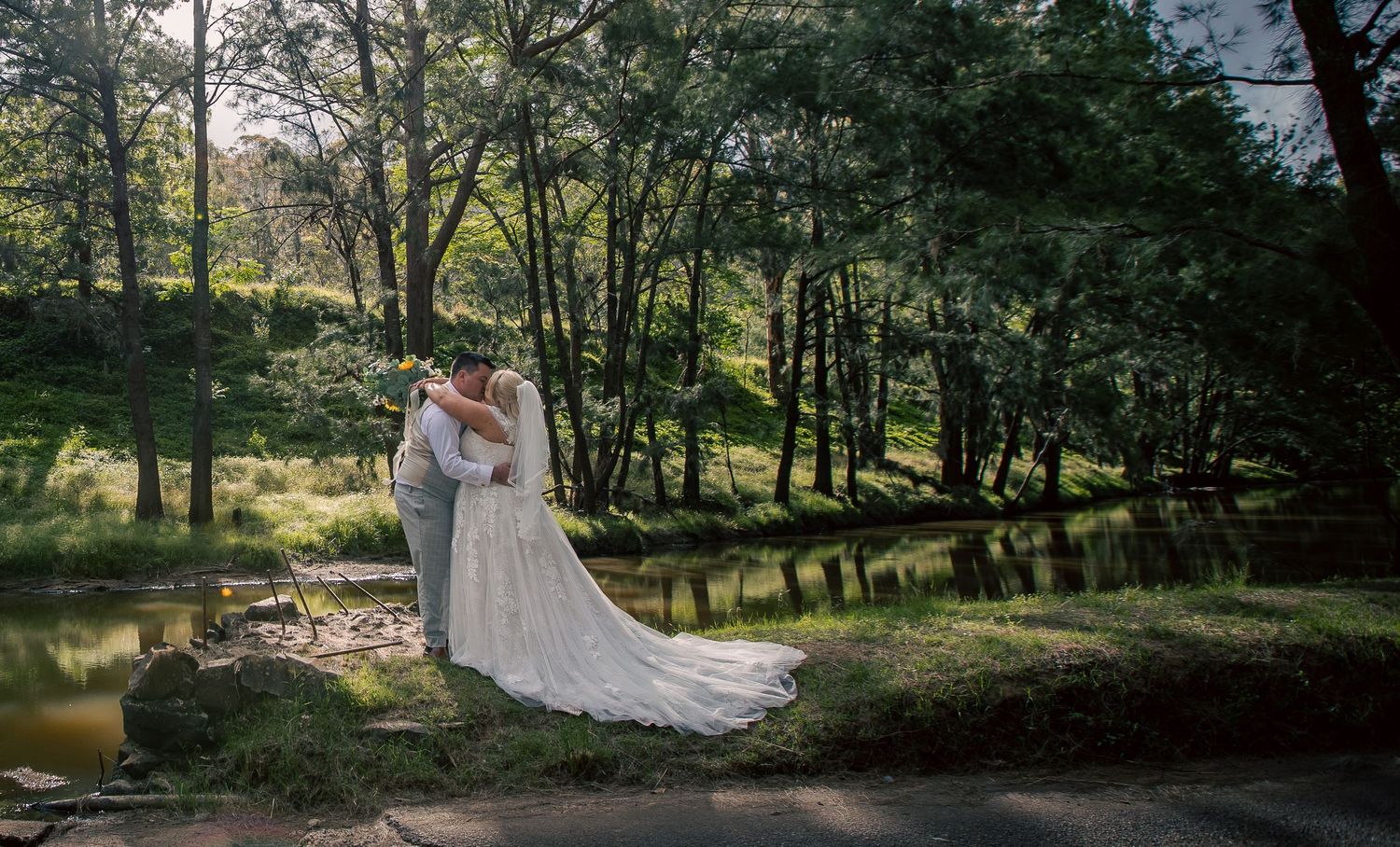 A couple shares an intimate moment in a serene woodland setting beside a tranquil pond at sunset.