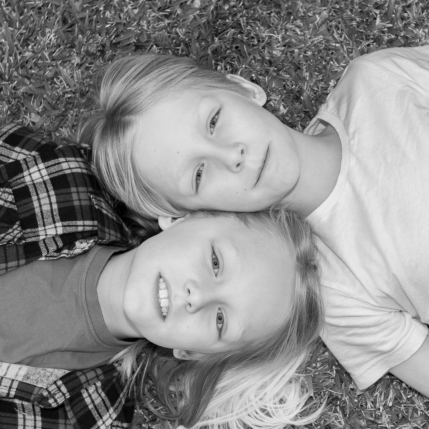 Close-up black and white portrait of two children lying head to head on grass.