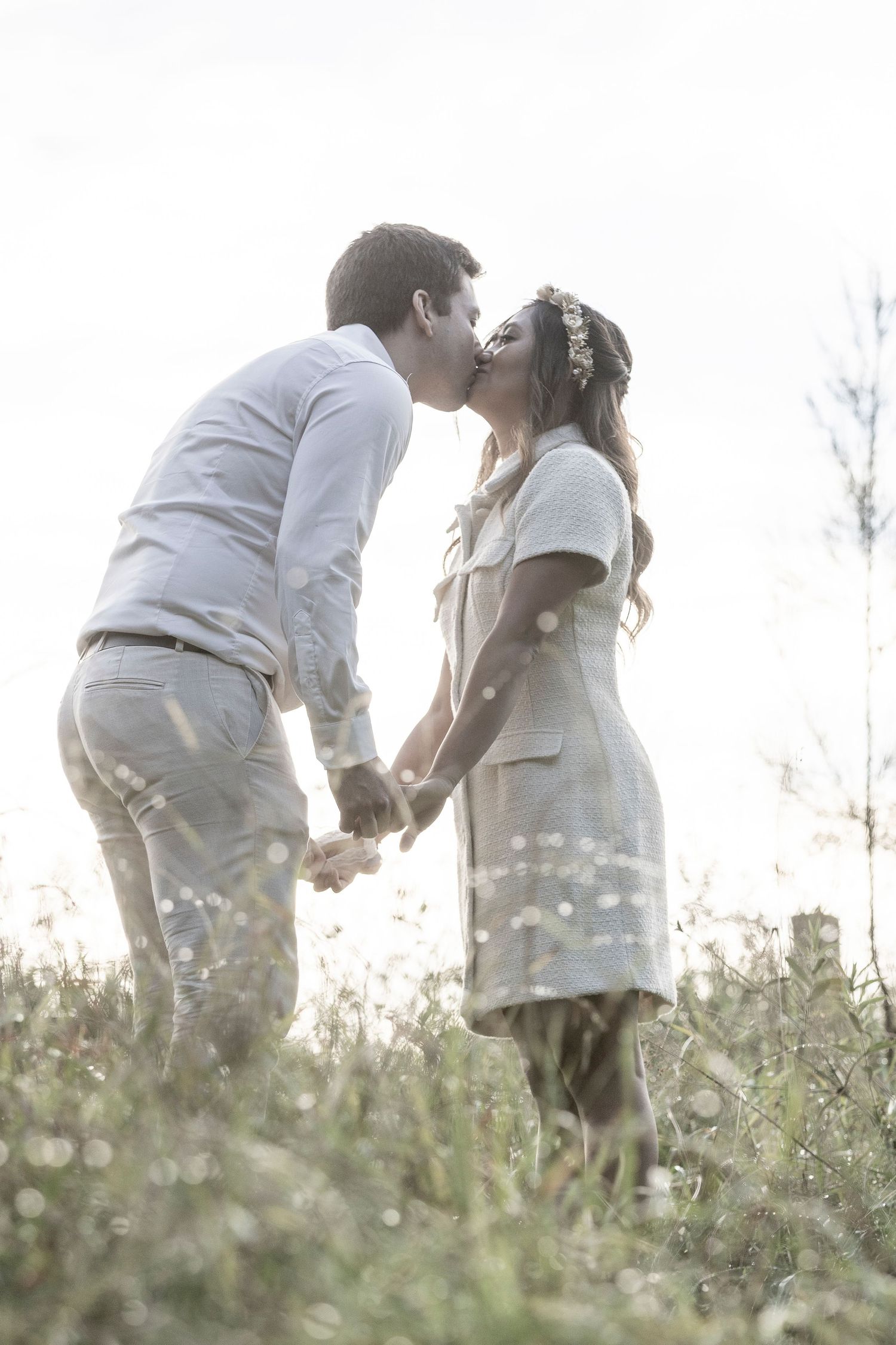 A romantic couple shares a tender kiss while holding hands in a sunlit meadow filled with wildflowers.