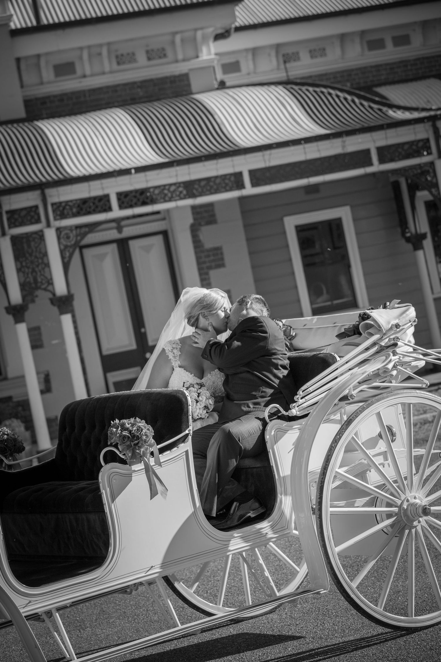 A black and white photograph of a couple leaning in close while seated in an ornate white horse-drawn carriage.