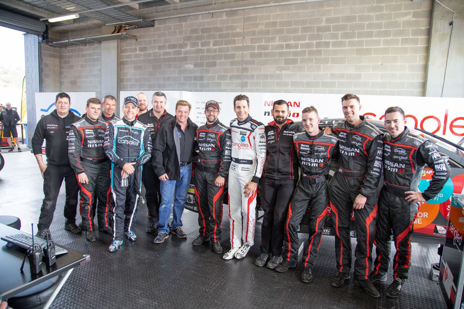Group of auto mechanics and service technicians standing in a line at a repair facility wearing dark uniforms. Chip Foose