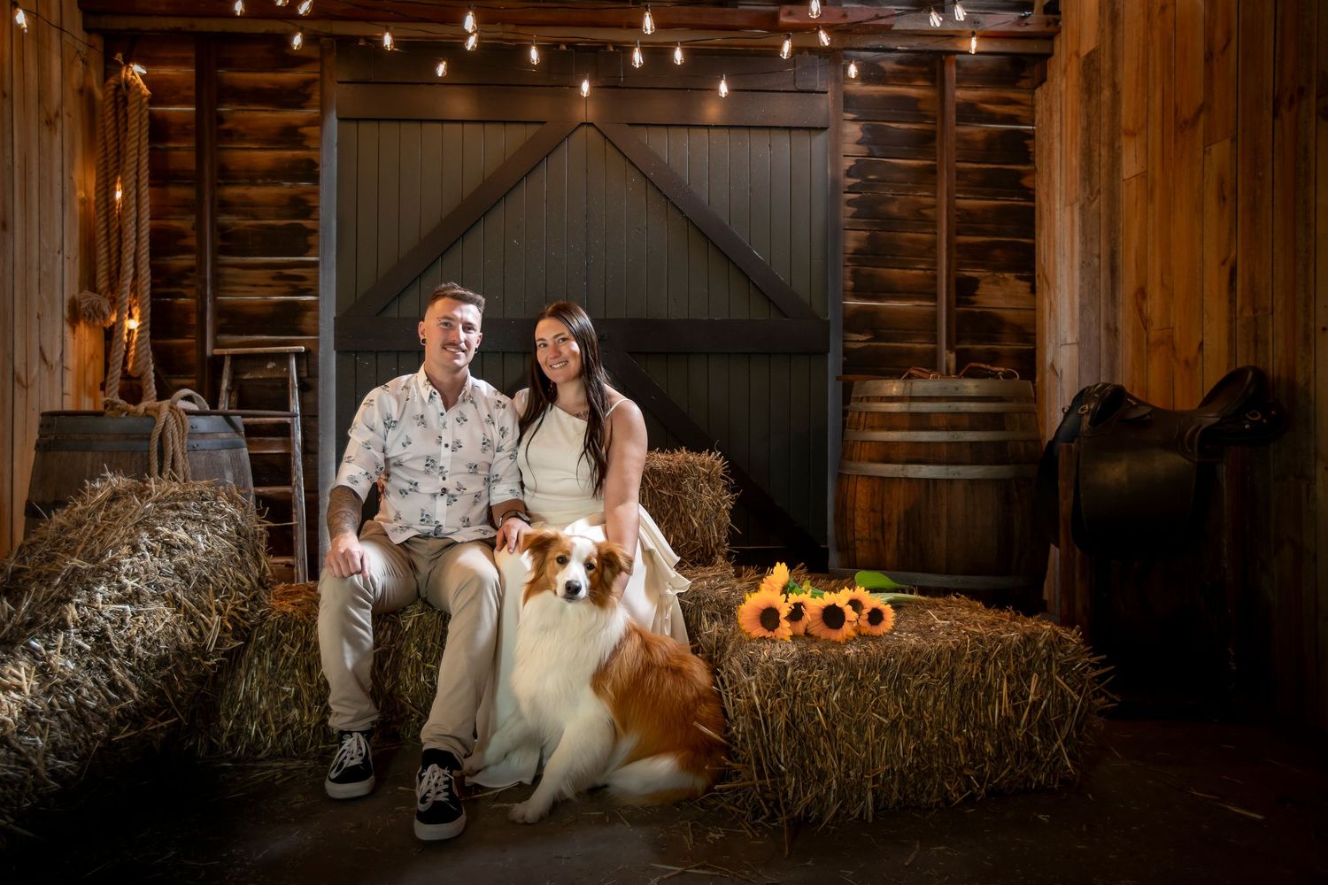 Couple and dog pose on hay bales under string lights in rustic barn setting.