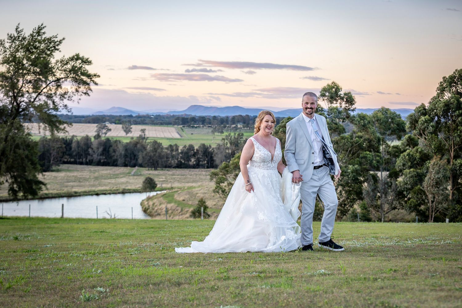 Couple in wedding attire walks together across scenic hillside meadow at sunset with mountains in background.