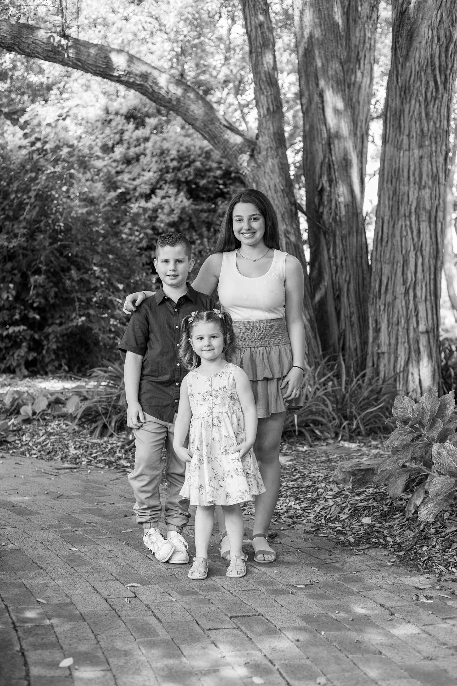 A series of four black and white outdoor photos showing a child in a floral dress on a brick path surrounded by trees.