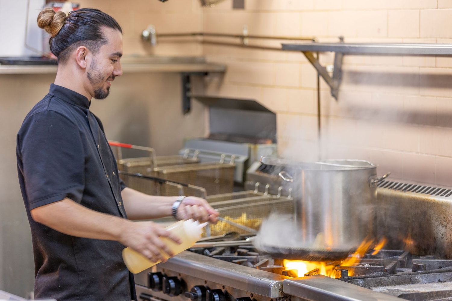 Professional cook in black uniform preparing food at a commercial kitchen stove with open flames.