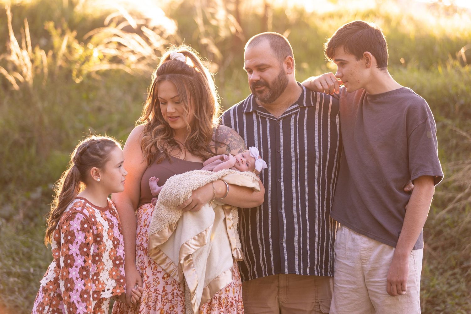 Family of five posing outdoors at golden hour, parents holding newborn baby surrounded by two children in a sunlit field.