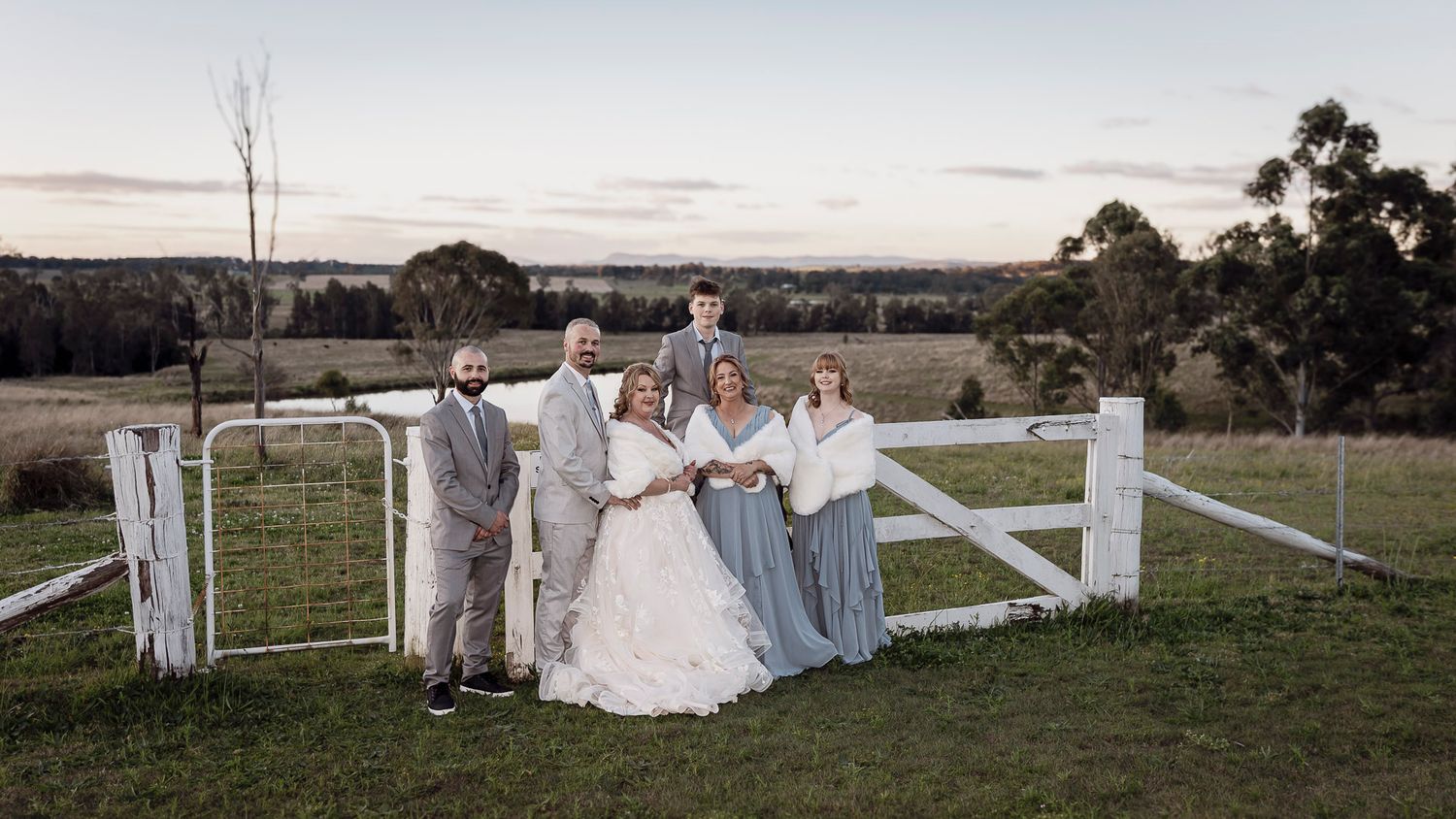Wedding couple poses in a rustic countryside setting with white fence and rolling hills in the background.