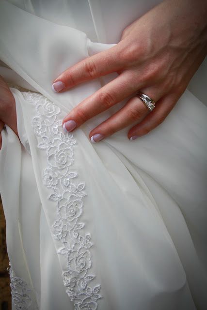 A manicured hand with a diamond ring rests on delicate white lace wedding dress fabric featuring floral embroidery.