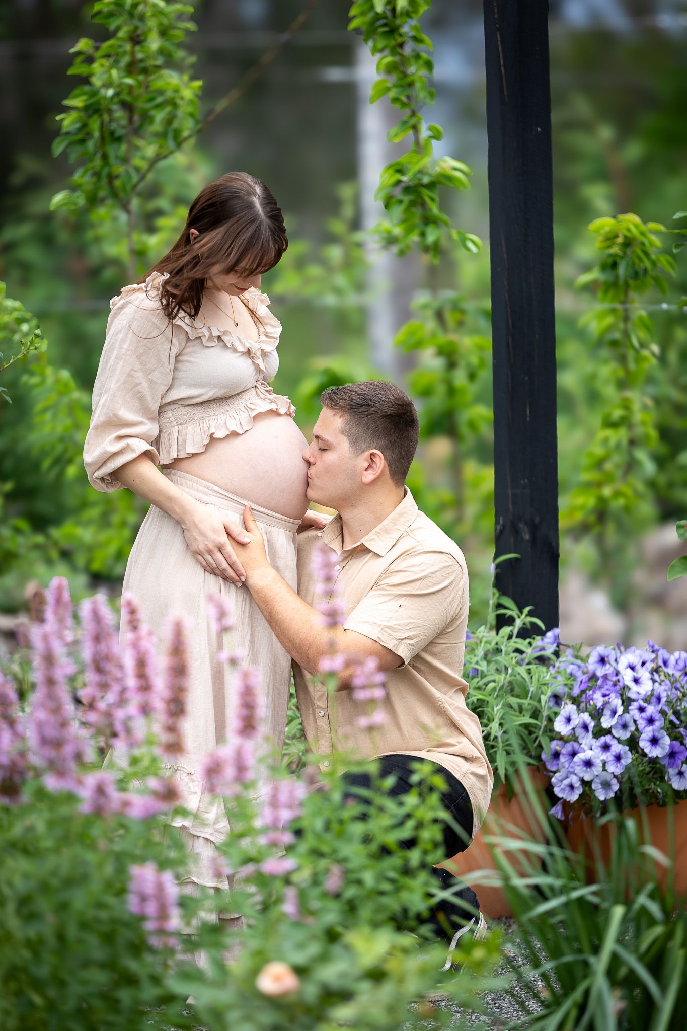 A romantic moment captured in a garden setting with pink and purple flowers blooming in the background for a maternity photos
