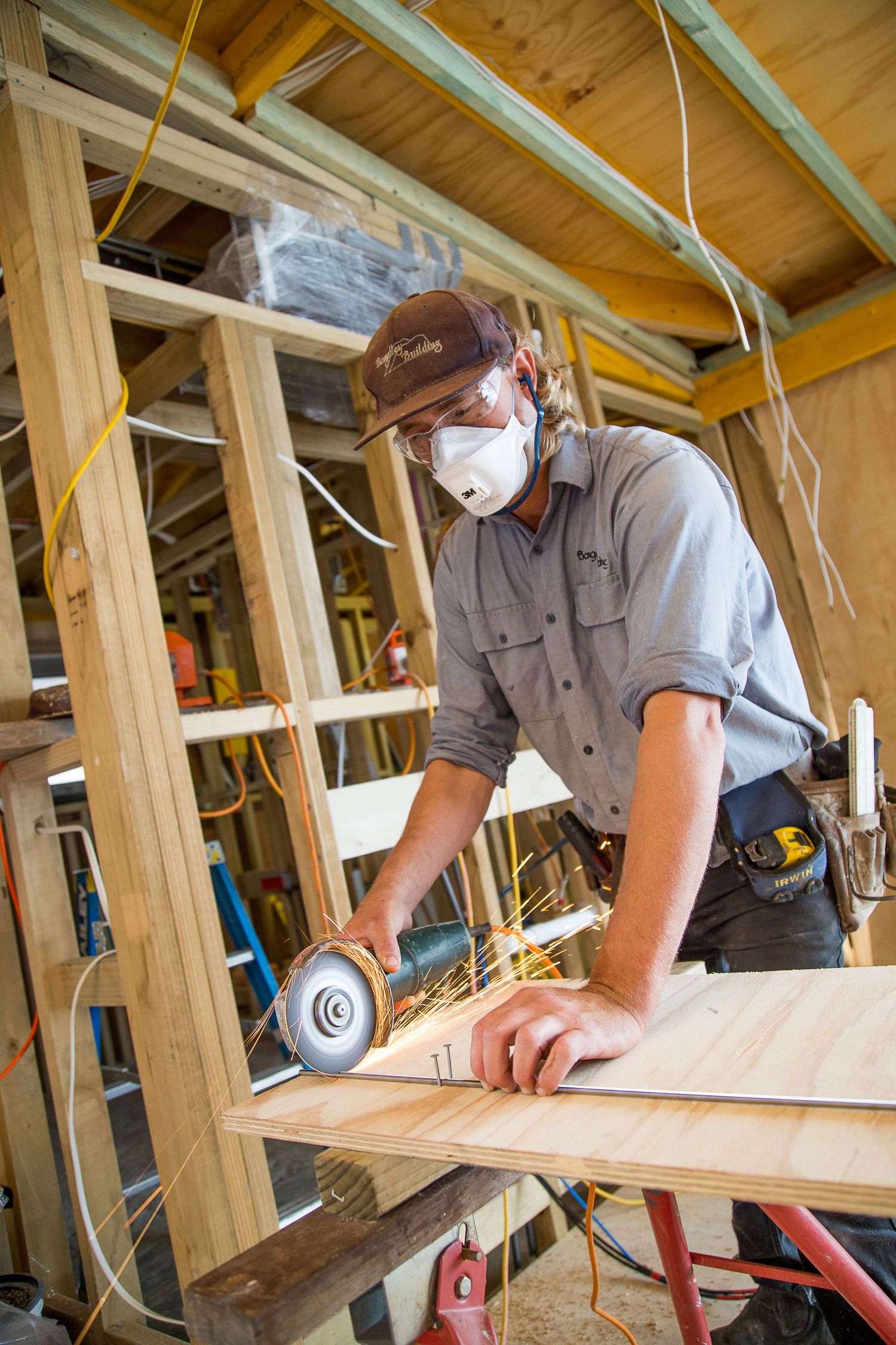 A carpenter wearing safety gear uses an angle grinder on wood in a workshop with exposed ceiling beams and tools.