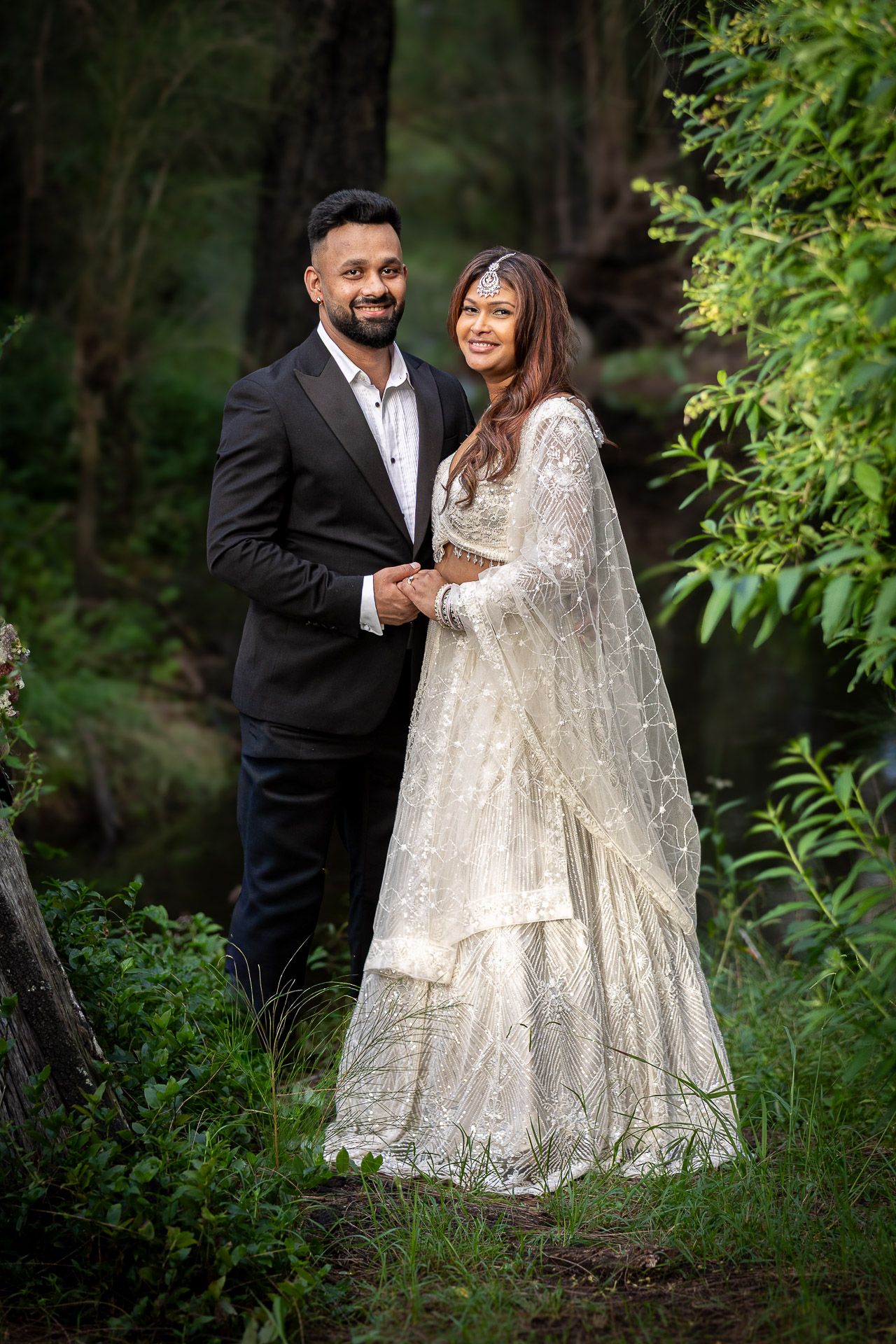 A couple in wedding attire stands together in a lush green forest setting for romantic outdoor portraits.