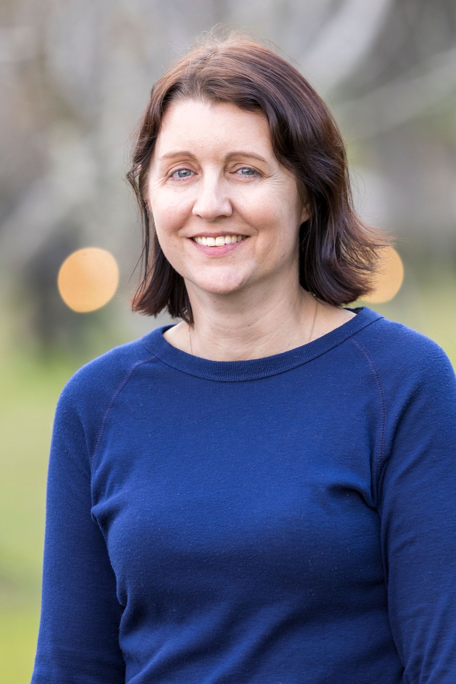 Person in navy blue sweater poses outdoors with soft bokeh lighting in background.