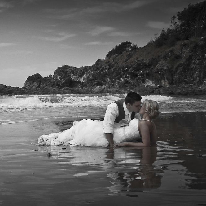 A romantic couple in formal wedding attire embracing in shallow ocean waters at sunset on a rocky beach in black and white.