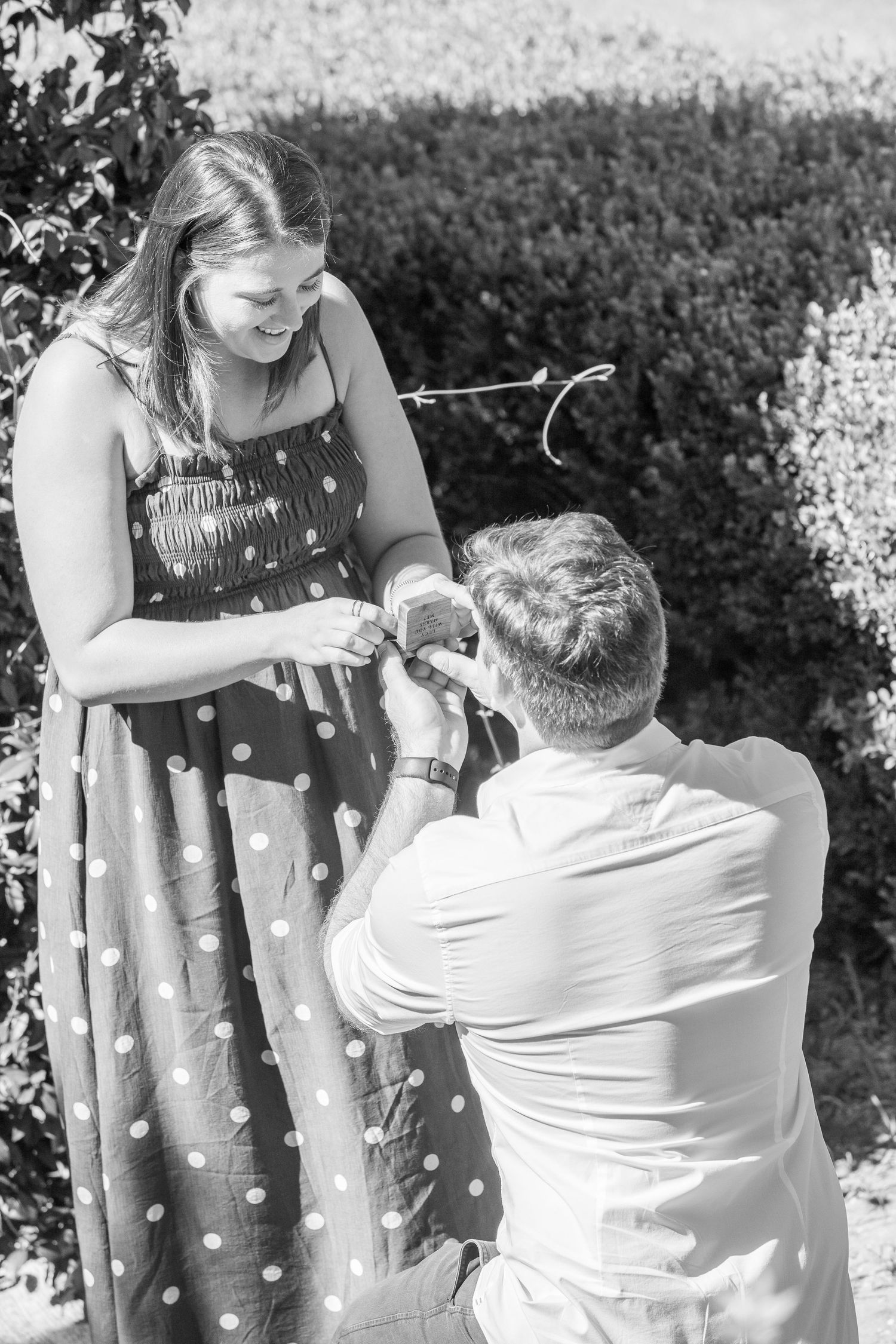 Black and white photo of a man on one knee proposing to a woman in a polka dot dress outdoors.