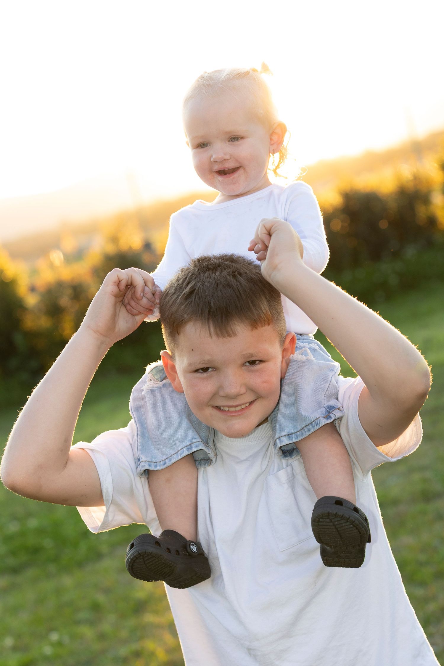 Joyful child in white shirt flexes muscles and smiles during golden hour sunset in grassy field.