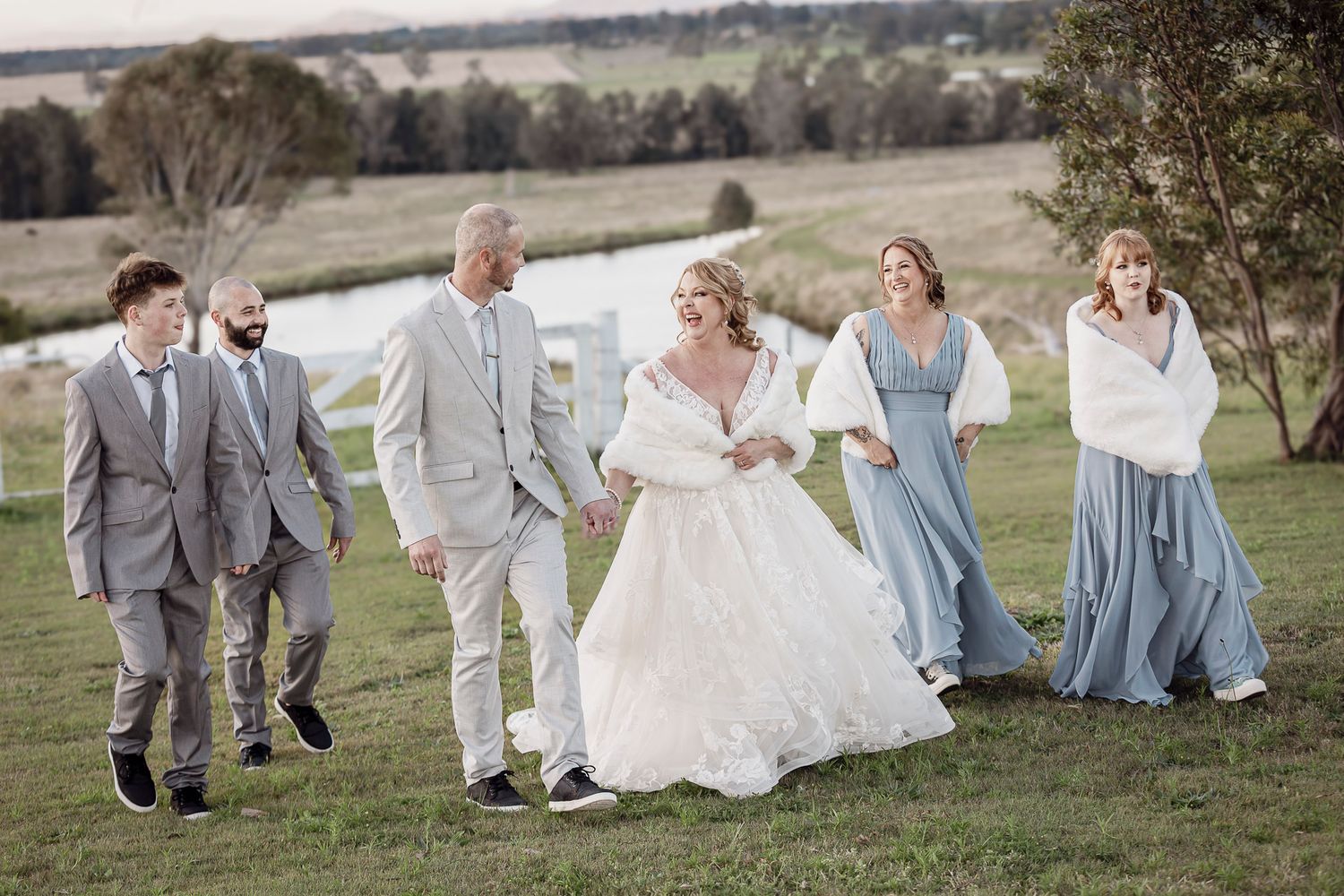 Wedding party in gray and blue attire playfully runs through a grassy field on a sunny day.