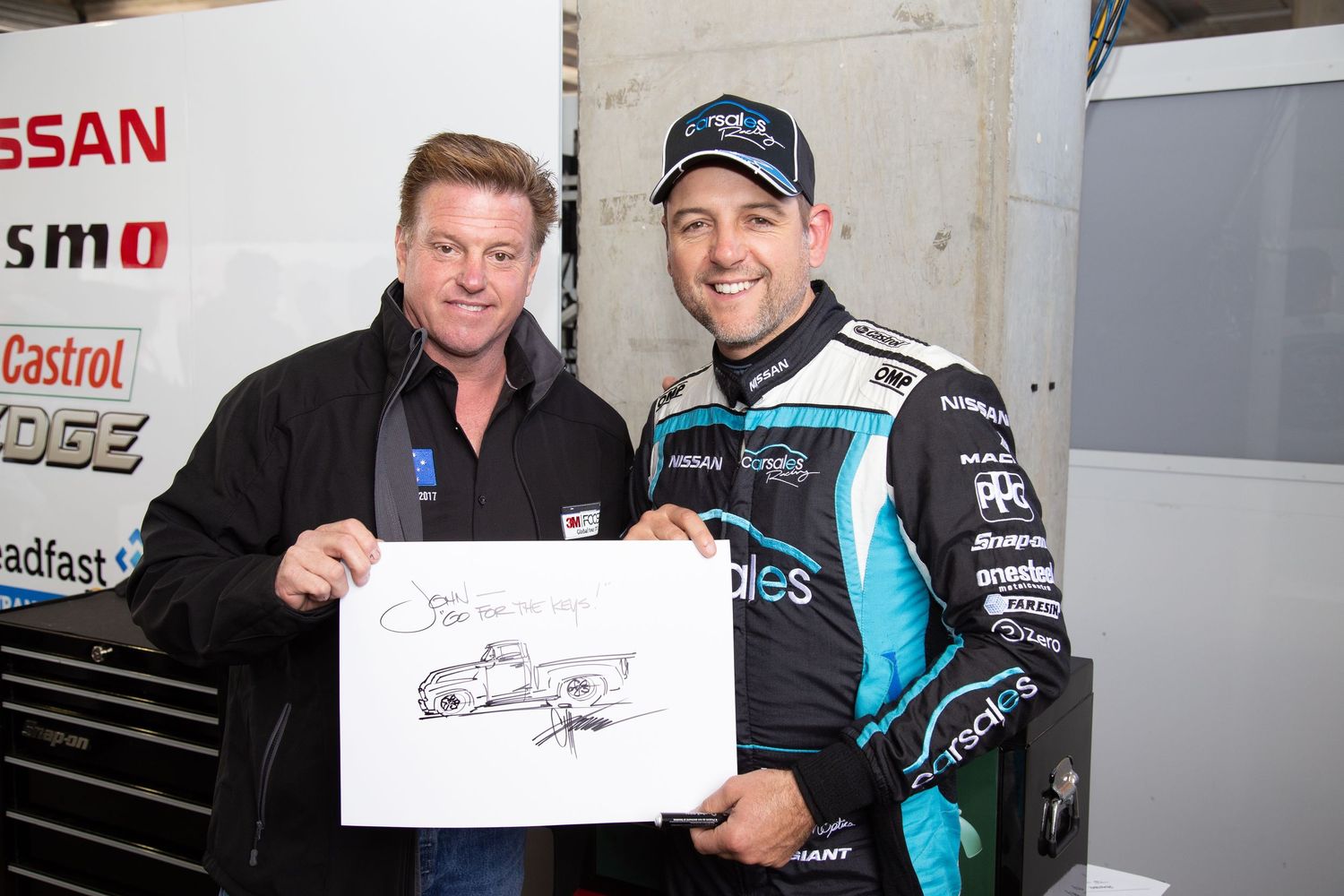 Two racing team members pose together in a pit garage with team merchandise and autographed memorabilia. Chip Foose