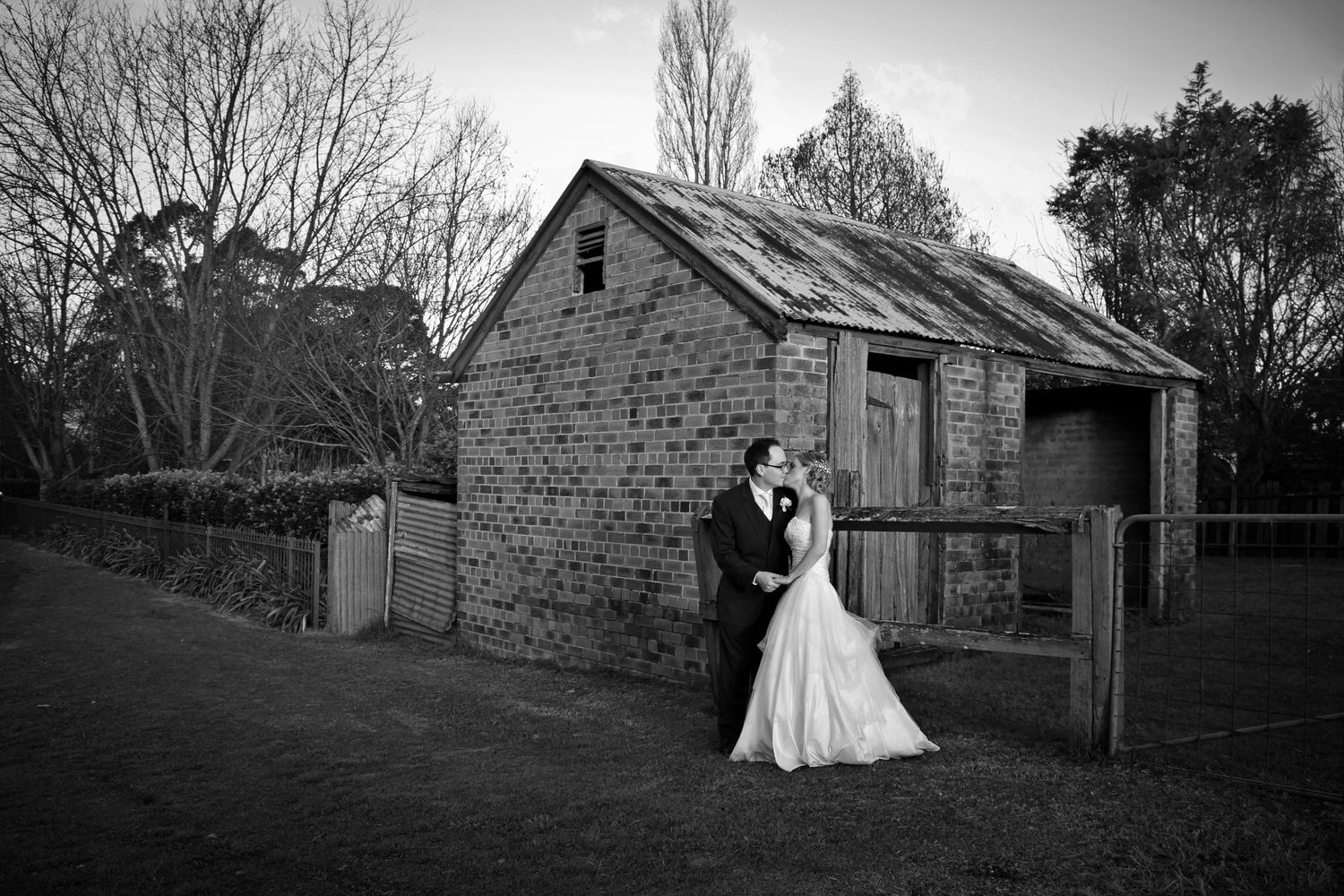 A romantic black and white photo of a couple embracing by a rustic stone barn in a pastoral setting.