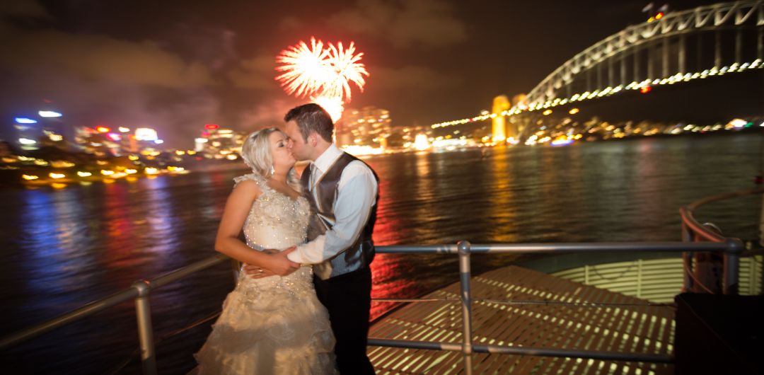 A couple shares a romantic moment by Sydney Harbour Bridge as fireworks light up the night sky.