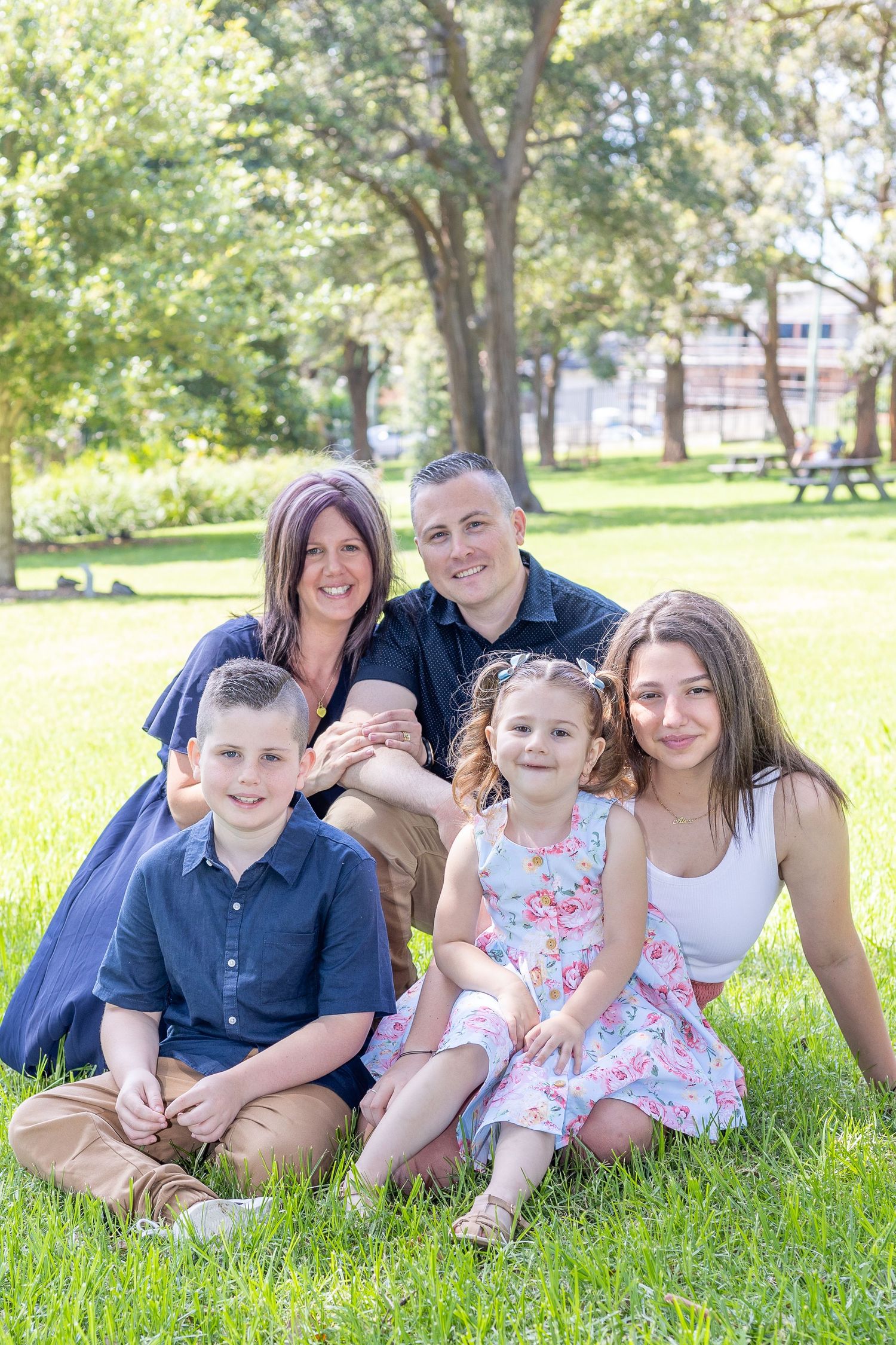 Photo link to Location Sessions. Family in blue and white outfits sitting together on grass in a sunny park with trees in background.