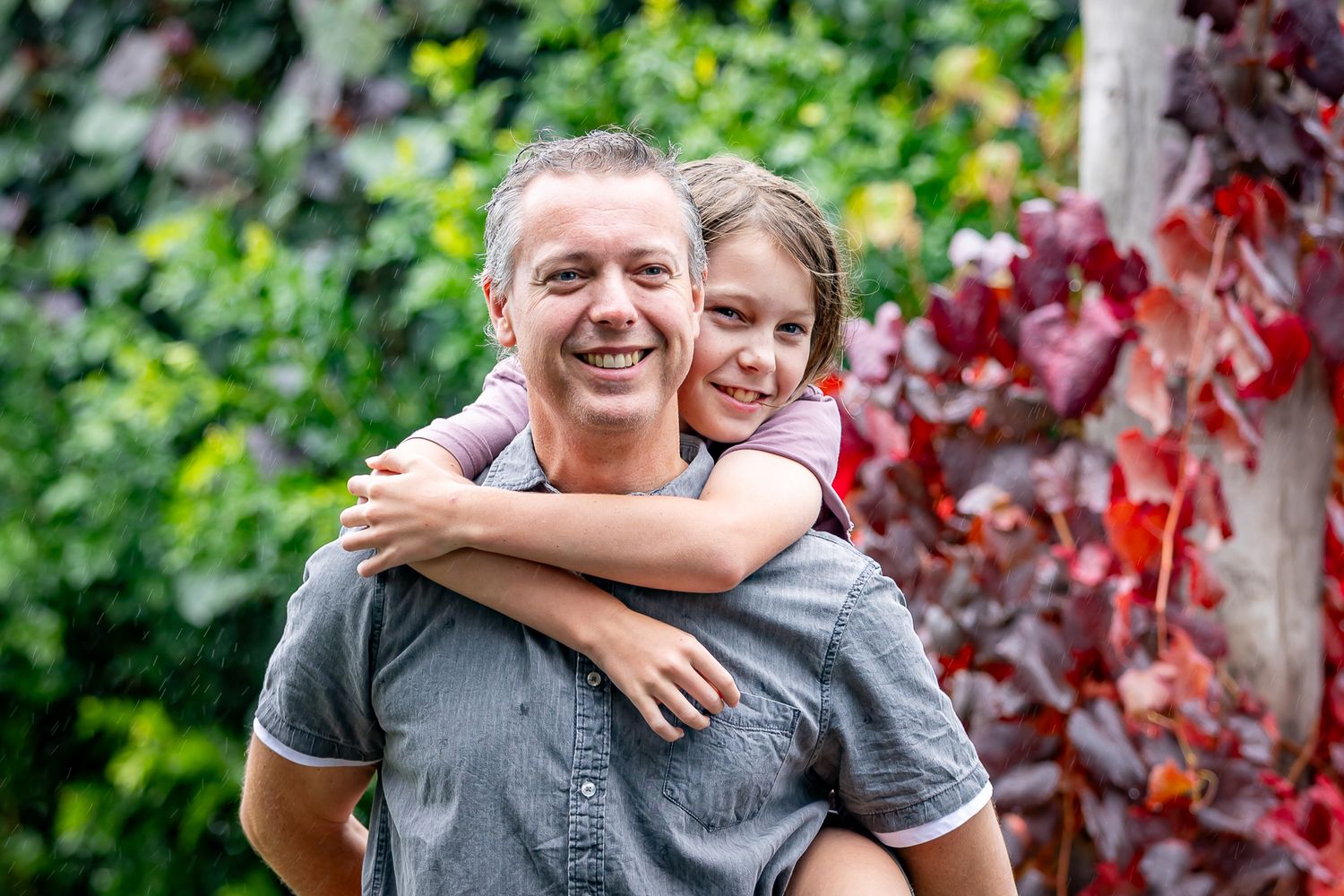A smiling man gives a happy young girl a piggyback ride in a lush garden with vibrant red autumn leaves.