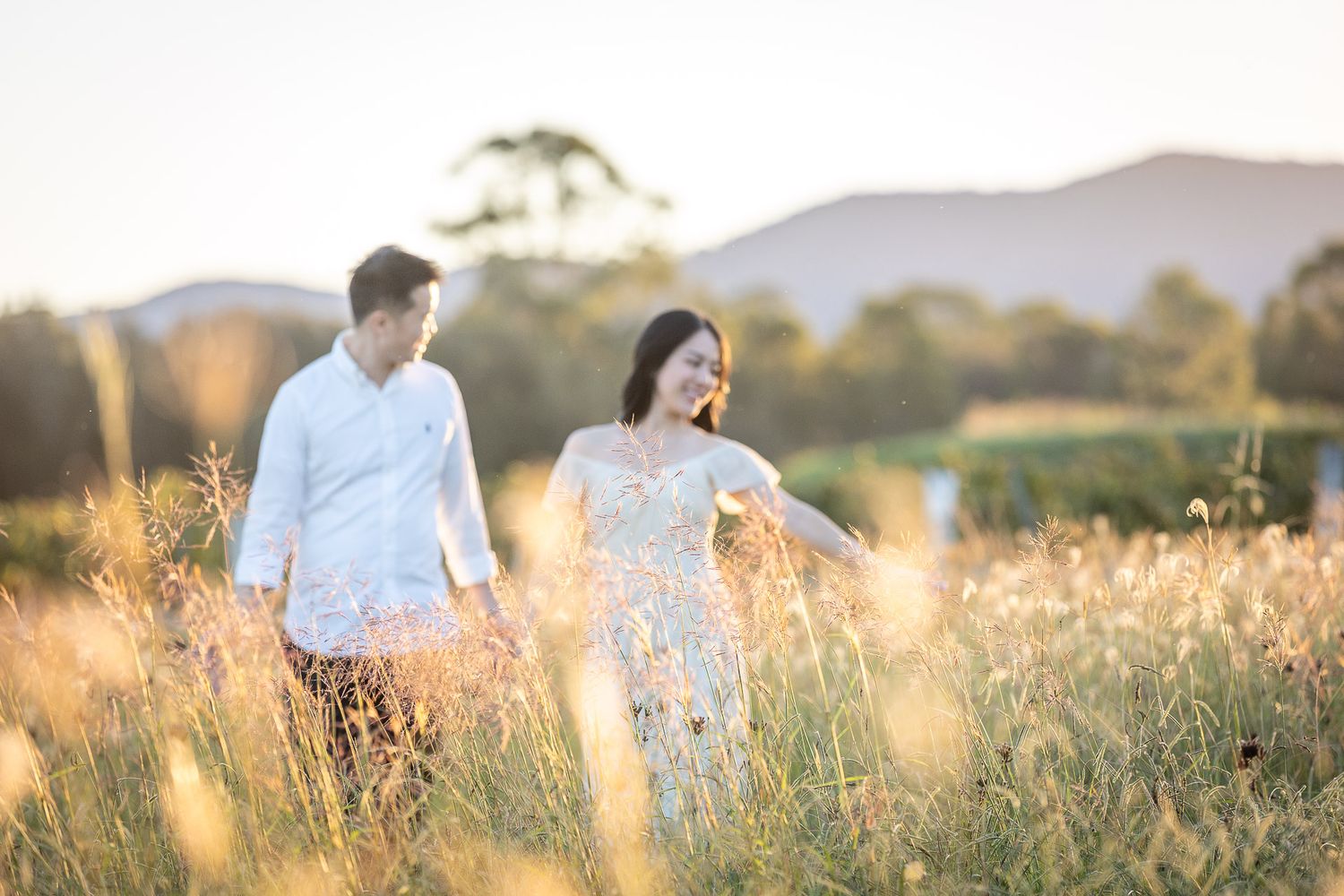 Couple walking hand in hand through golden meadow at sunset with mountains in background.