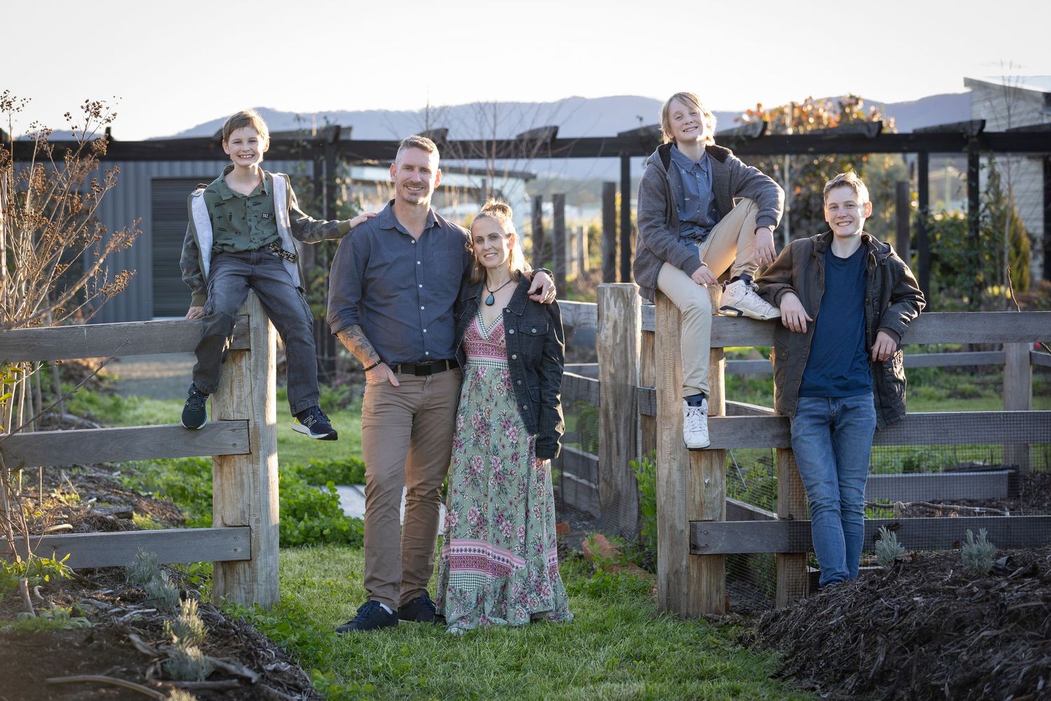 Group of people posing casually at wooden gates in a rural garden setting during golden hour lighting.