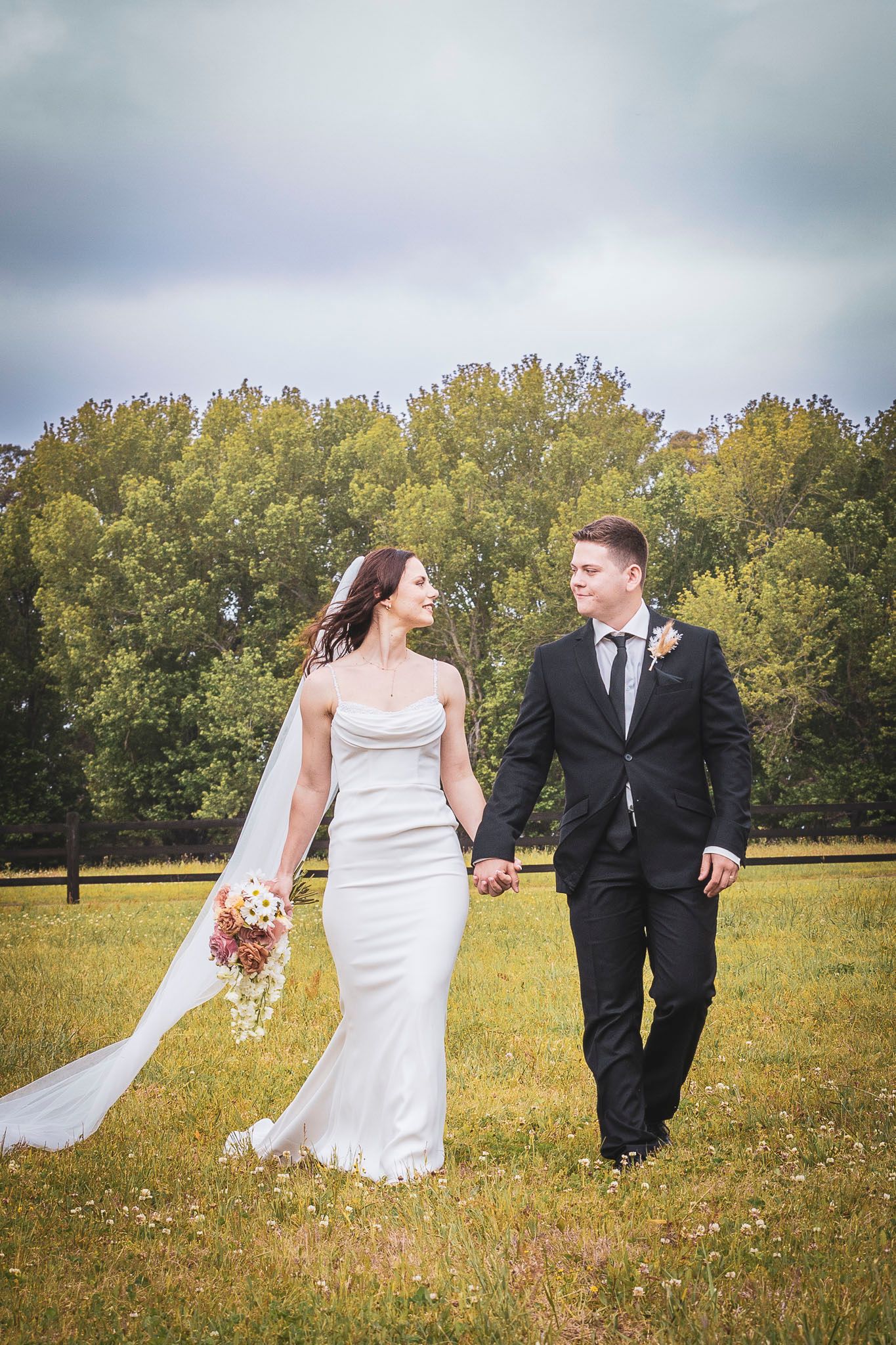 A bride and groom walk hand in hand through a scenic autumn field with golden grass and trees in the background.