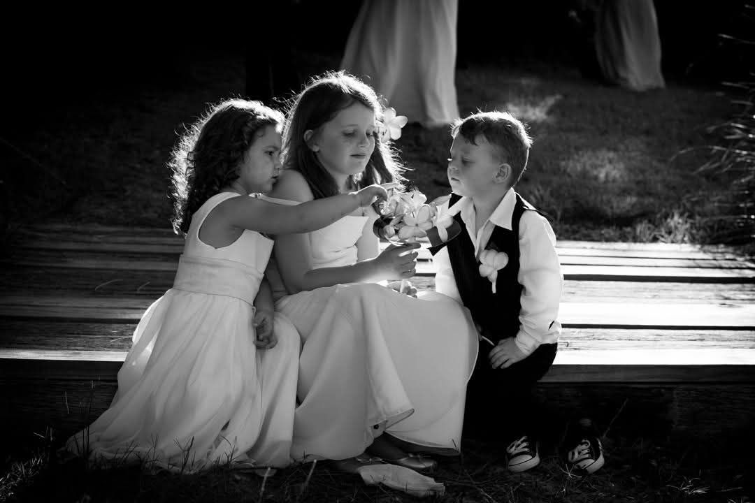 Black and white photo of three young children in formal attire laughing together outdoors in natural light.