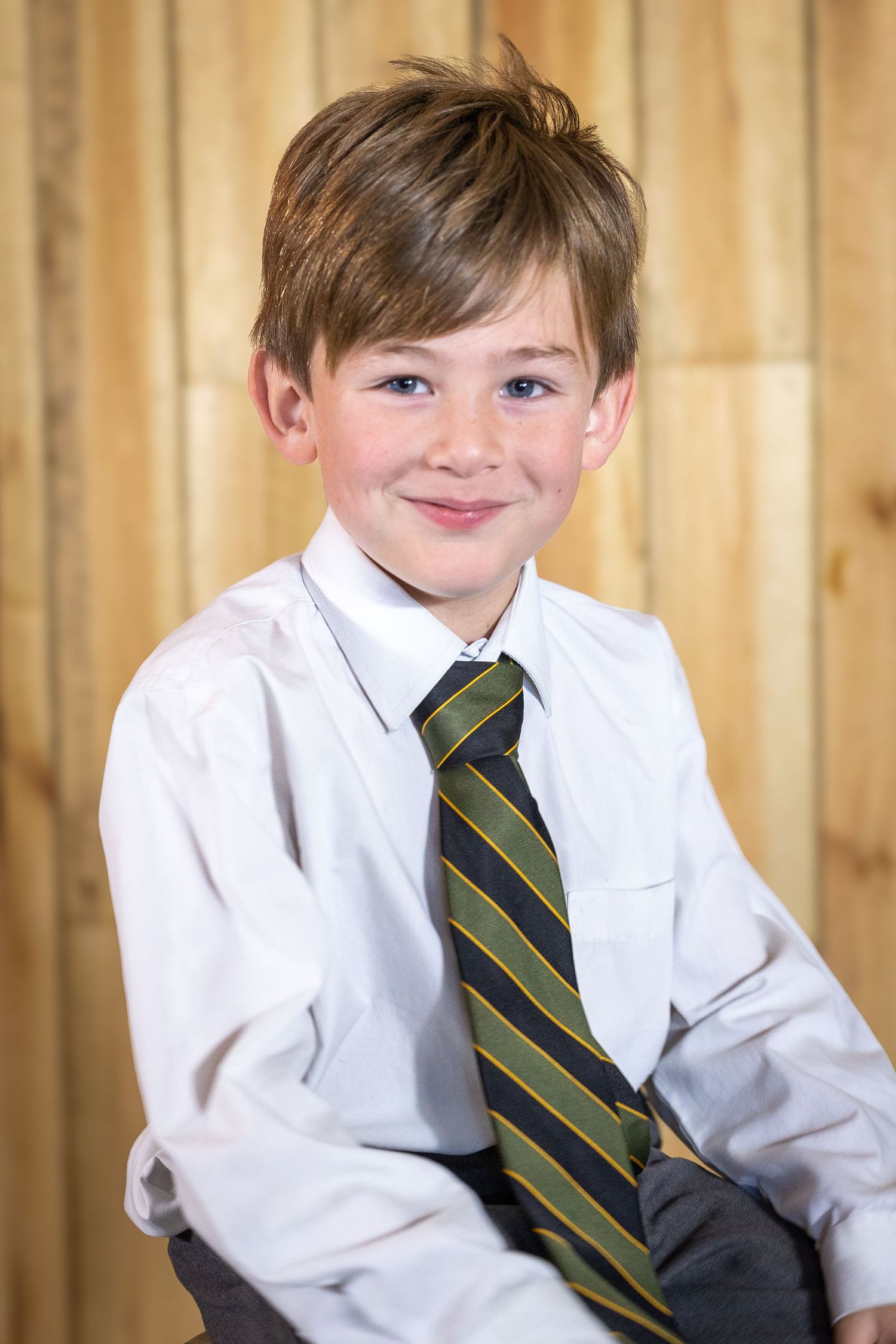Young boy in white shirt and striped tie smiling for a school portrait photo against wooden background.