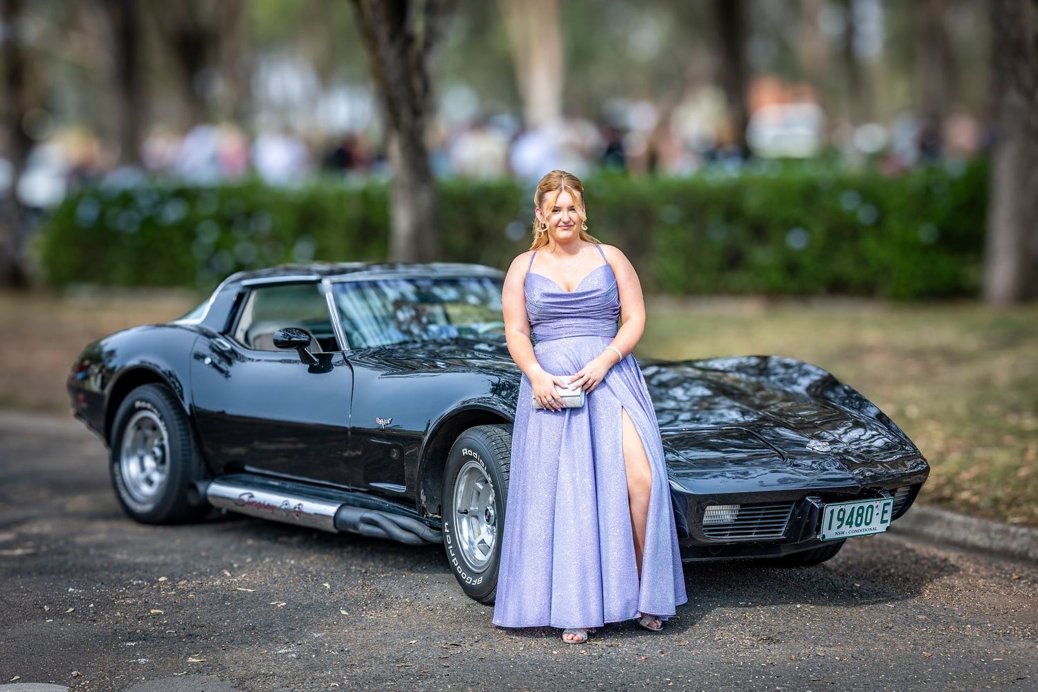 Young woman in lavender prom gown posing beside a classic black Corvette sports car outdoors.