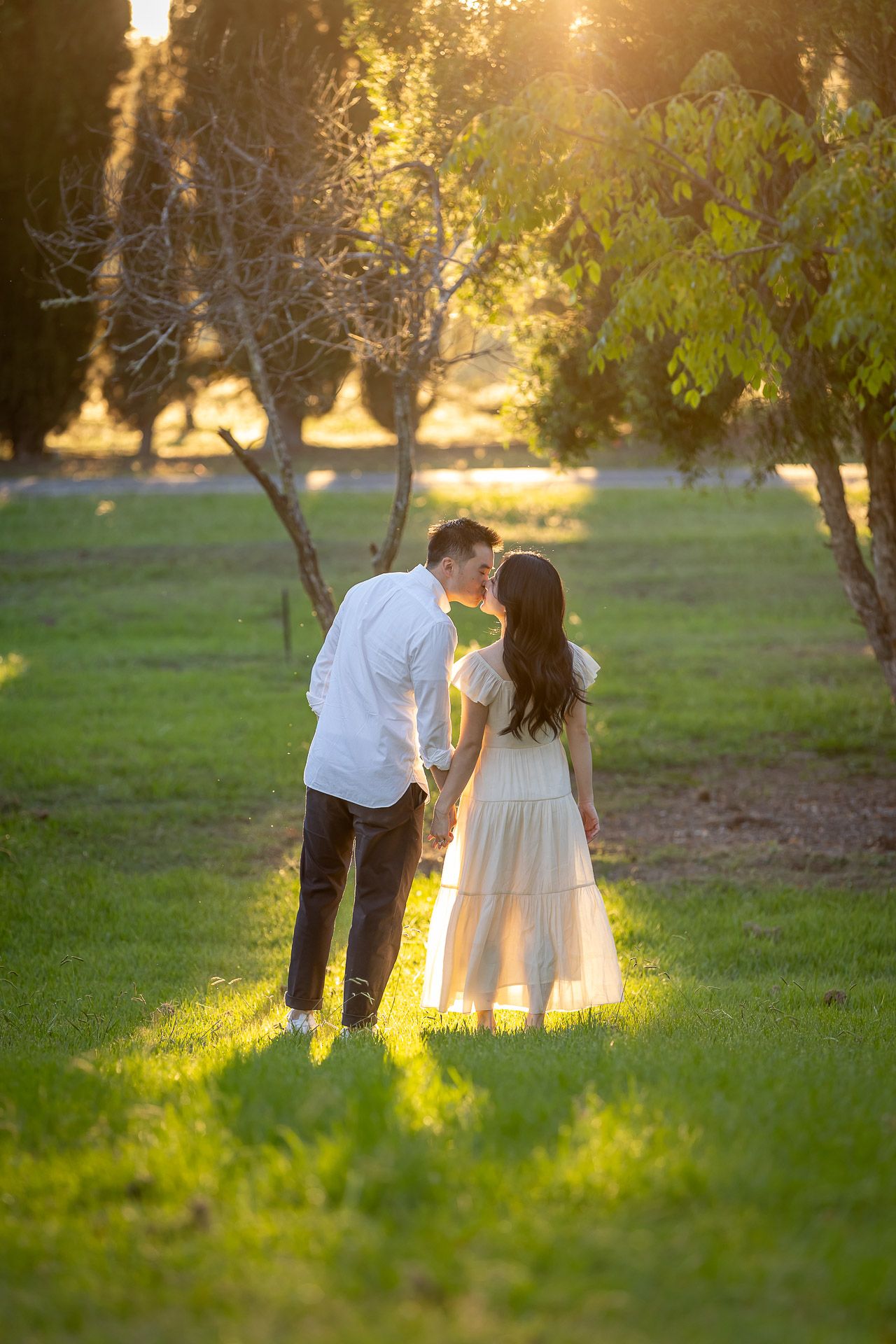 Couple sharing a romantic kiss in a sunlit park, man in white shirt and woman in white dress holding hands.