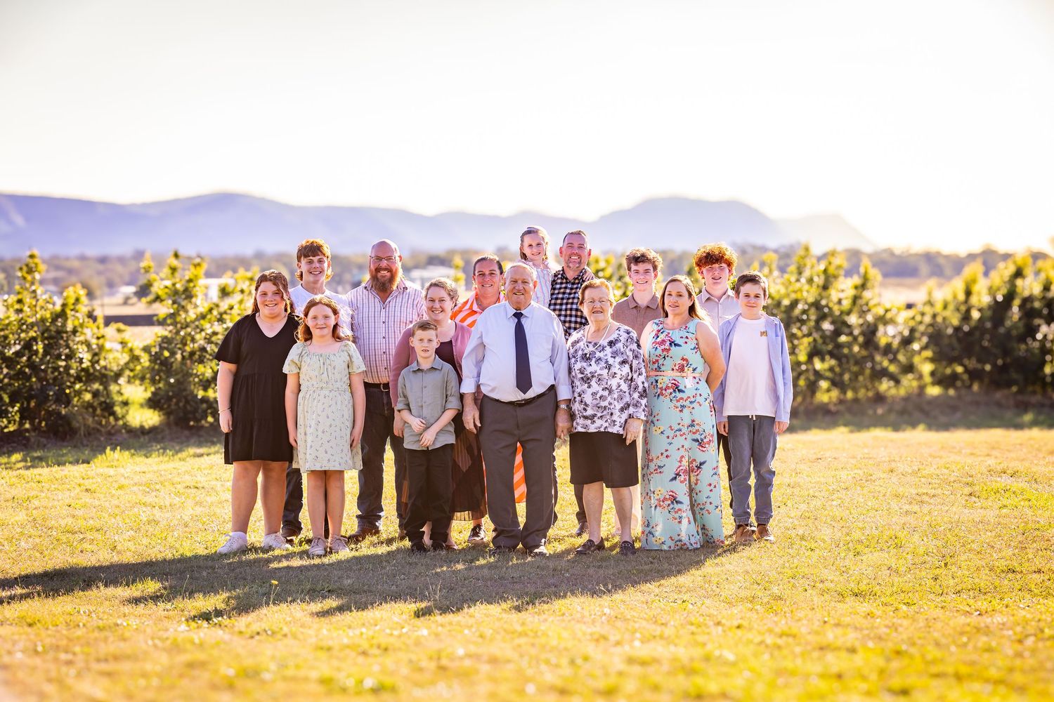 Large family group portrait outdoors at golden hour with mountains and greenery in background.