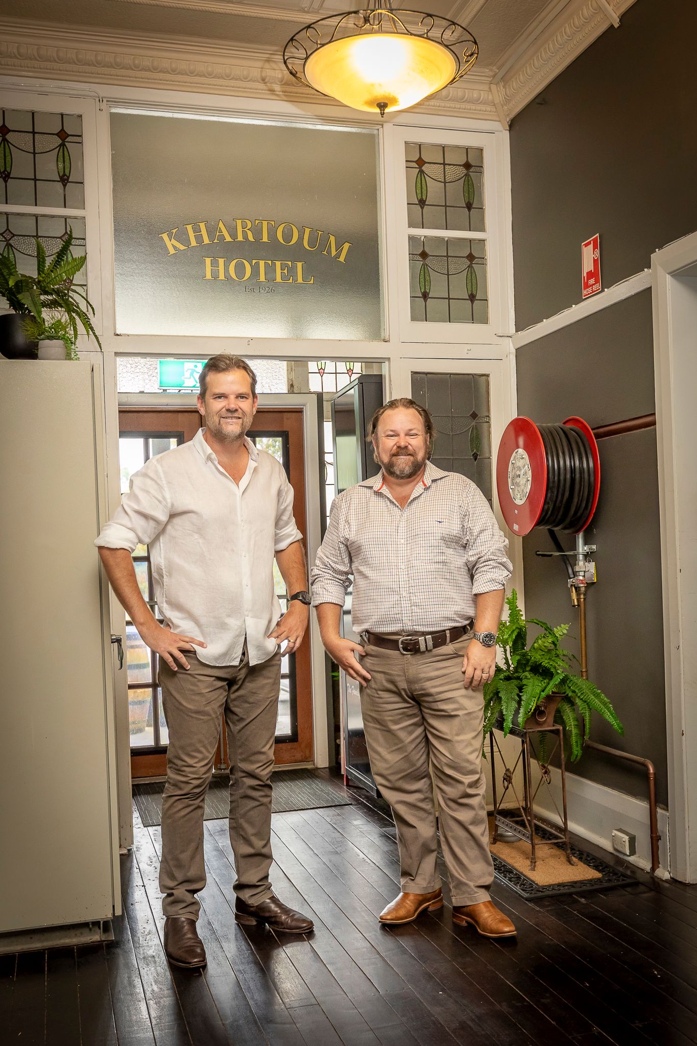 Two people in matching uniforms stand in a hotel hallway with dark wood floors and vintage decor.