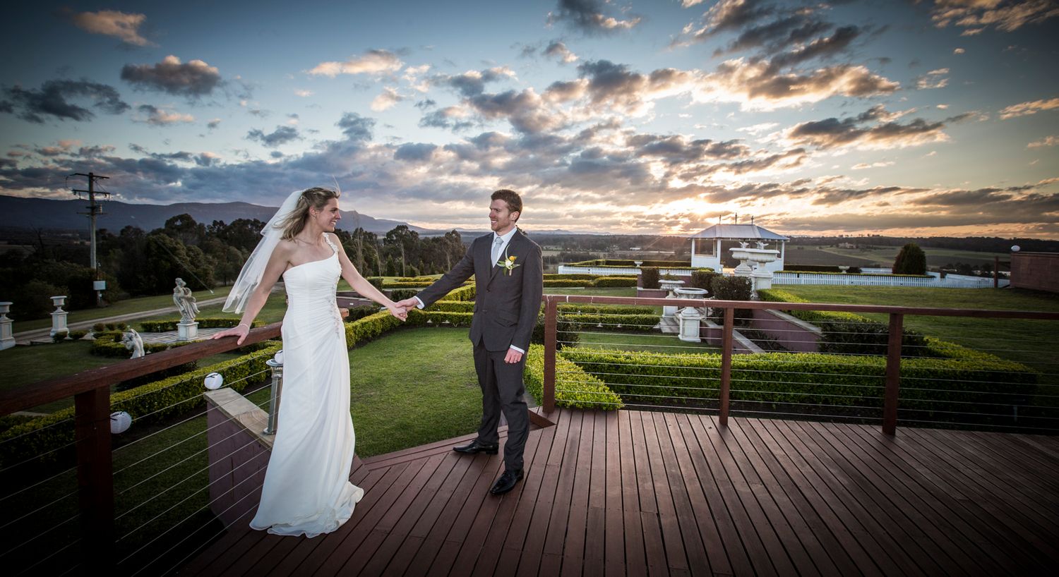 A couple stands on an outdoor deck overlooking a scenic landscape with a dramatic sunset and clouds in the background.